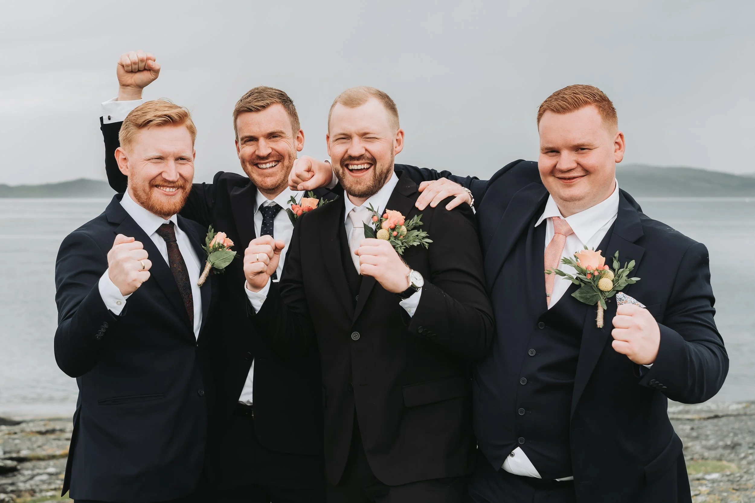 Fire fornøyde menn i dress med blomsterknapper står sammen ved stranden og jubler.