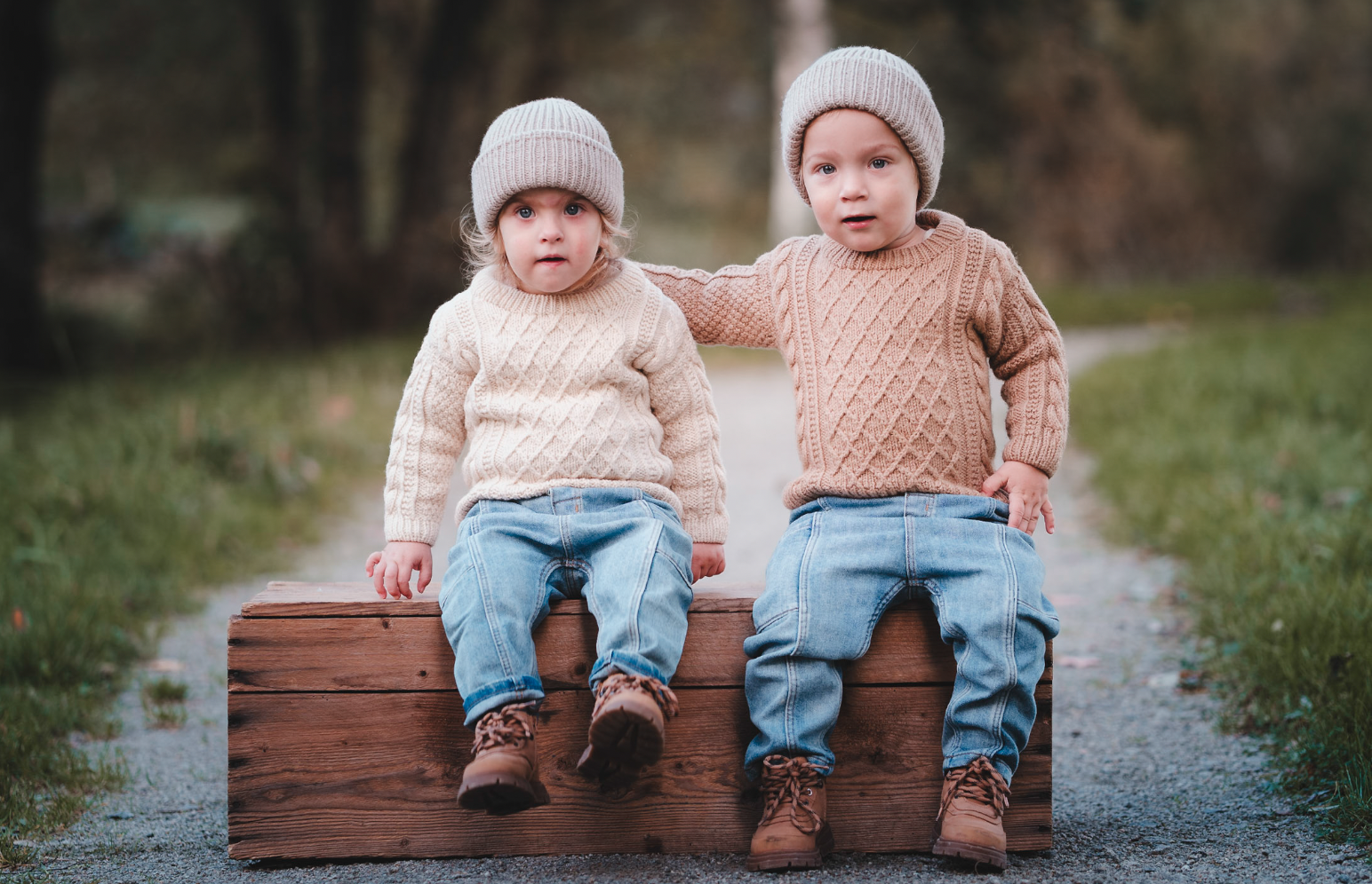 To små barn, en gutt og ei jente, sitter på en trebenk i en park med høstfarger, iført strikkegensere, chili og bukser.