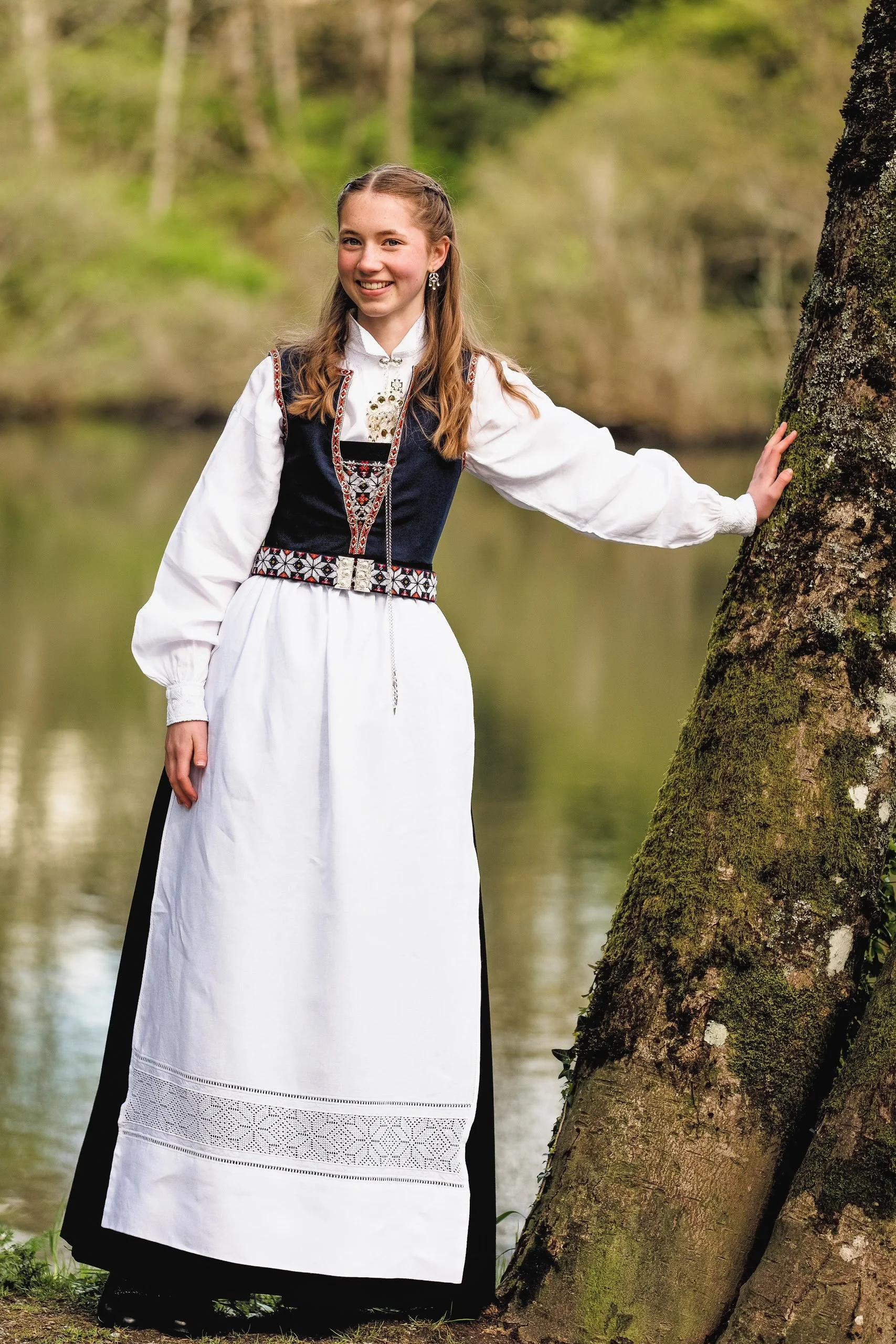 En ung kvinne i tradisjonell norsk bunad står ved en trestamme ved vann, smilende og ser mot kamera.