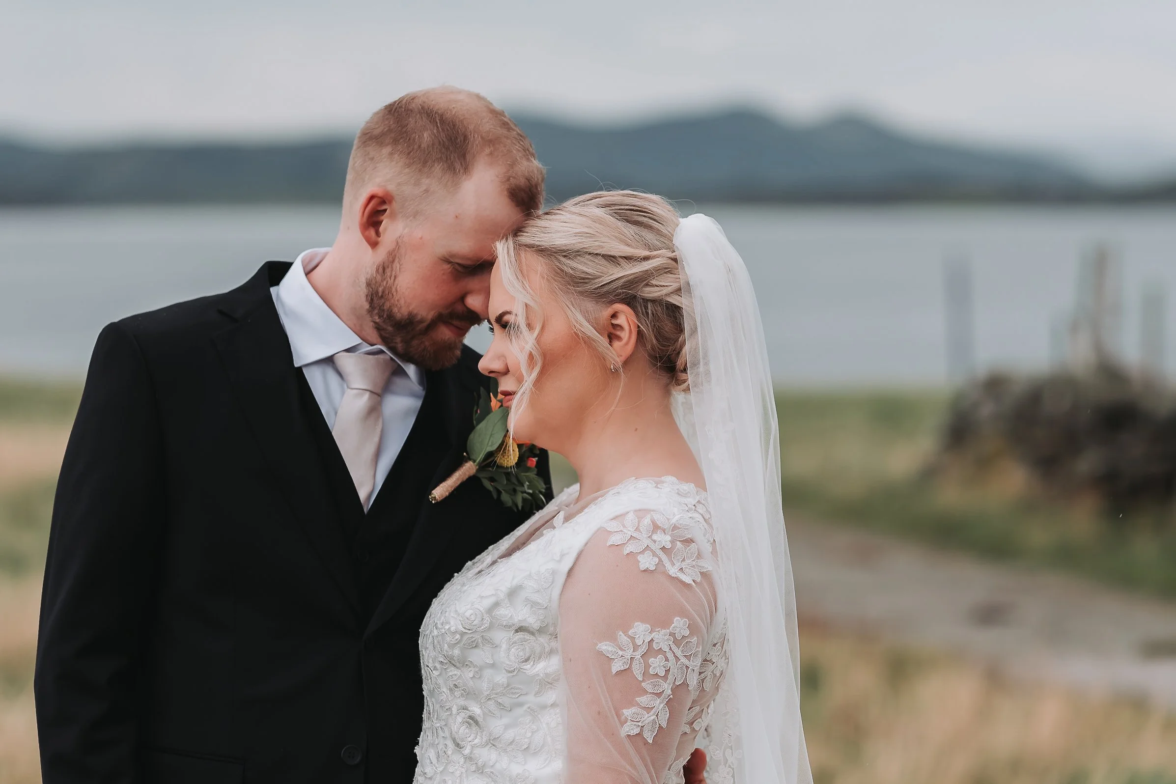 Nybrudepar i svart dress og hvit blondekjole omfavner hverandre foran fjell og vann