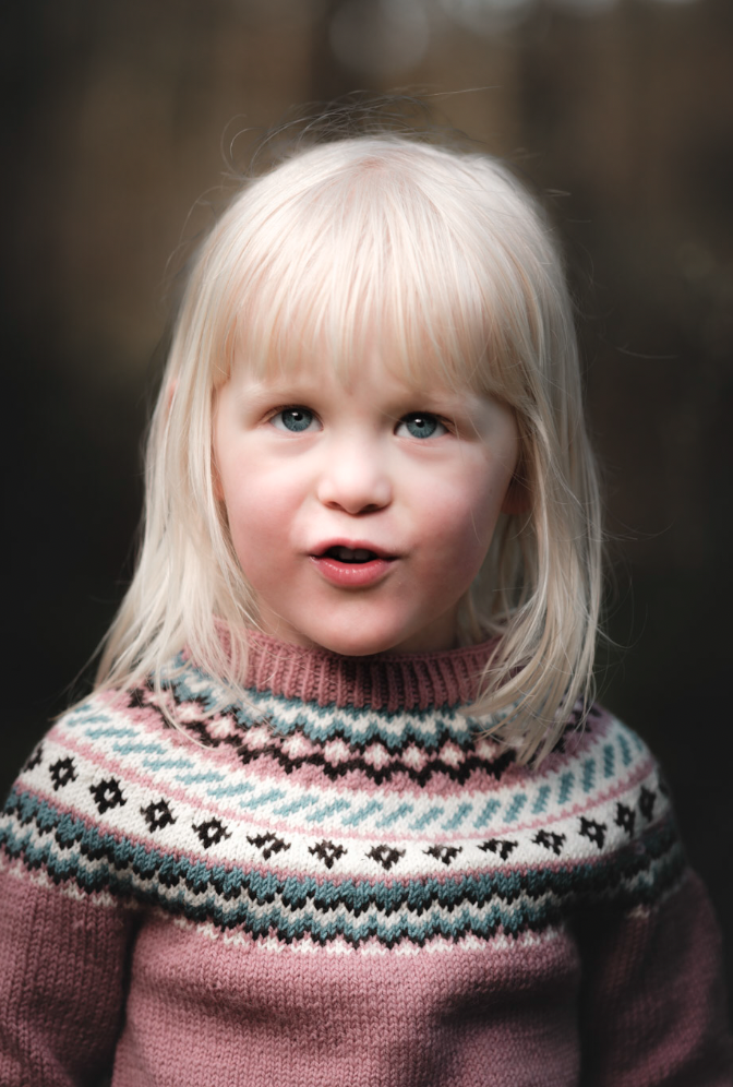 Liten jente med blond hår, blå øyne, iført en mønstret genser, står utendørs.