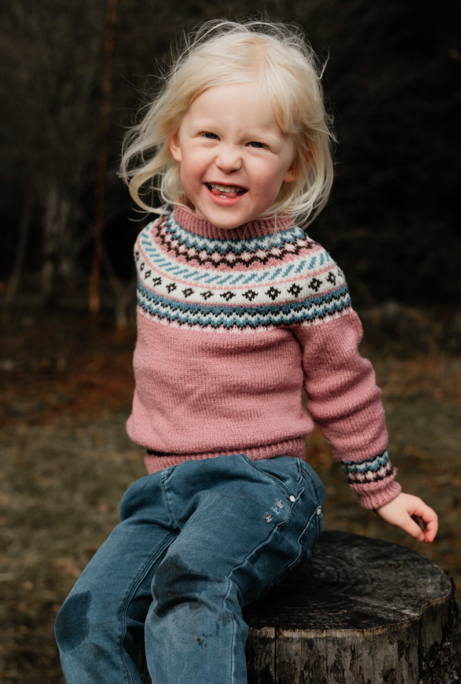 Liten jente med blondt hår smilende på en utendørs trestubbe, iført rosa genser med mønster og blå bukser, i en naturlig skogsomgivelse.