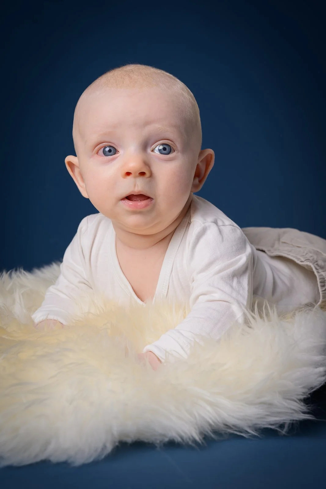Smilende baby i hvit body som sitter på et mykt, hvitt teppe mot mørkeblå bakgrunn.
