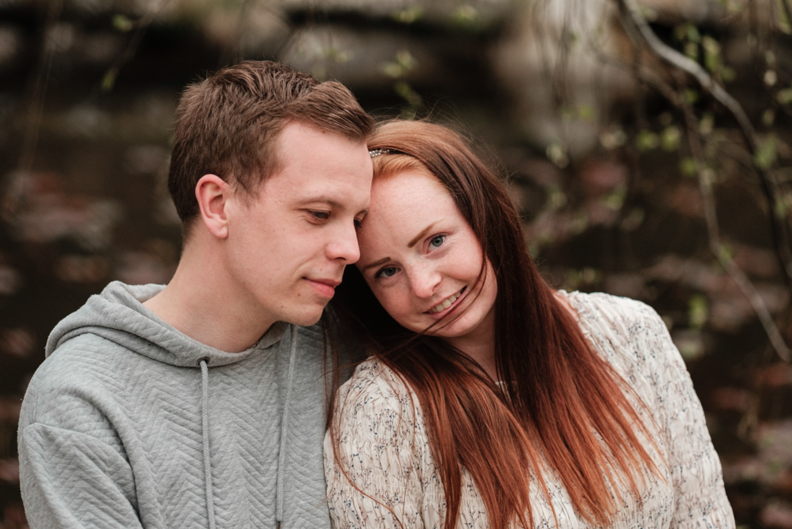 Et ungt par og ei ung jente som står tett sammen utendørs, med et nøysomt uttrykk, foran en bakgrunn av mørkegrønne blader og grener.