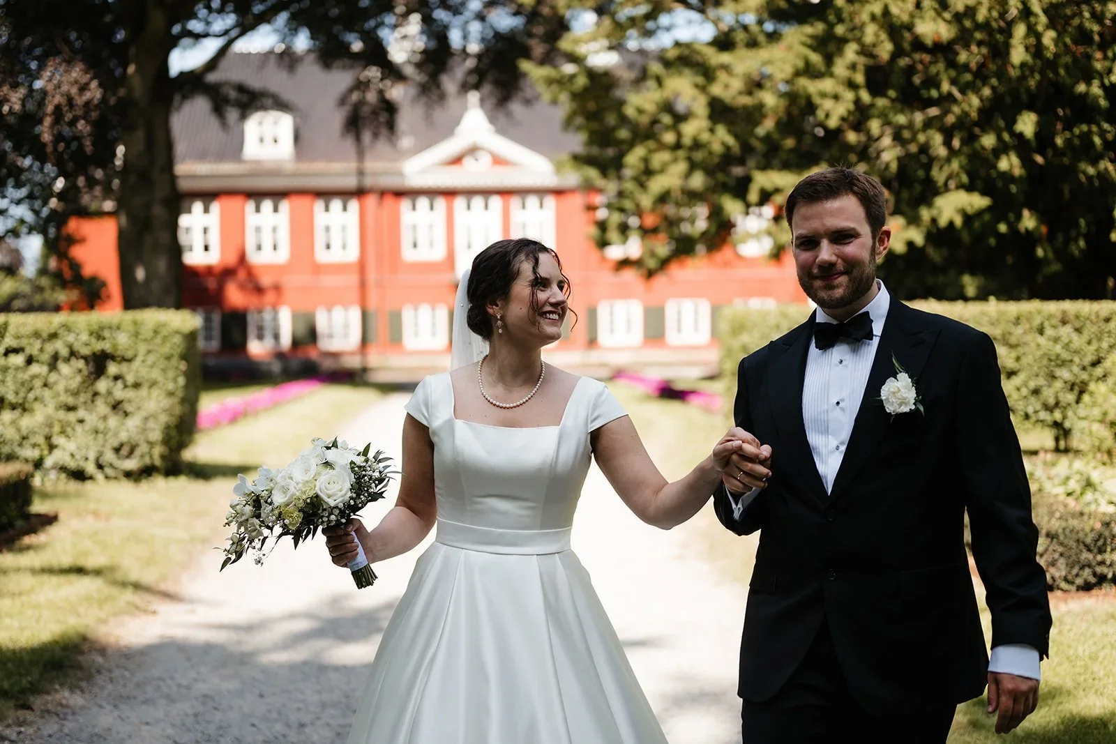 Brudepar går hånd i hånd foran et rødt herskapshus i en park
