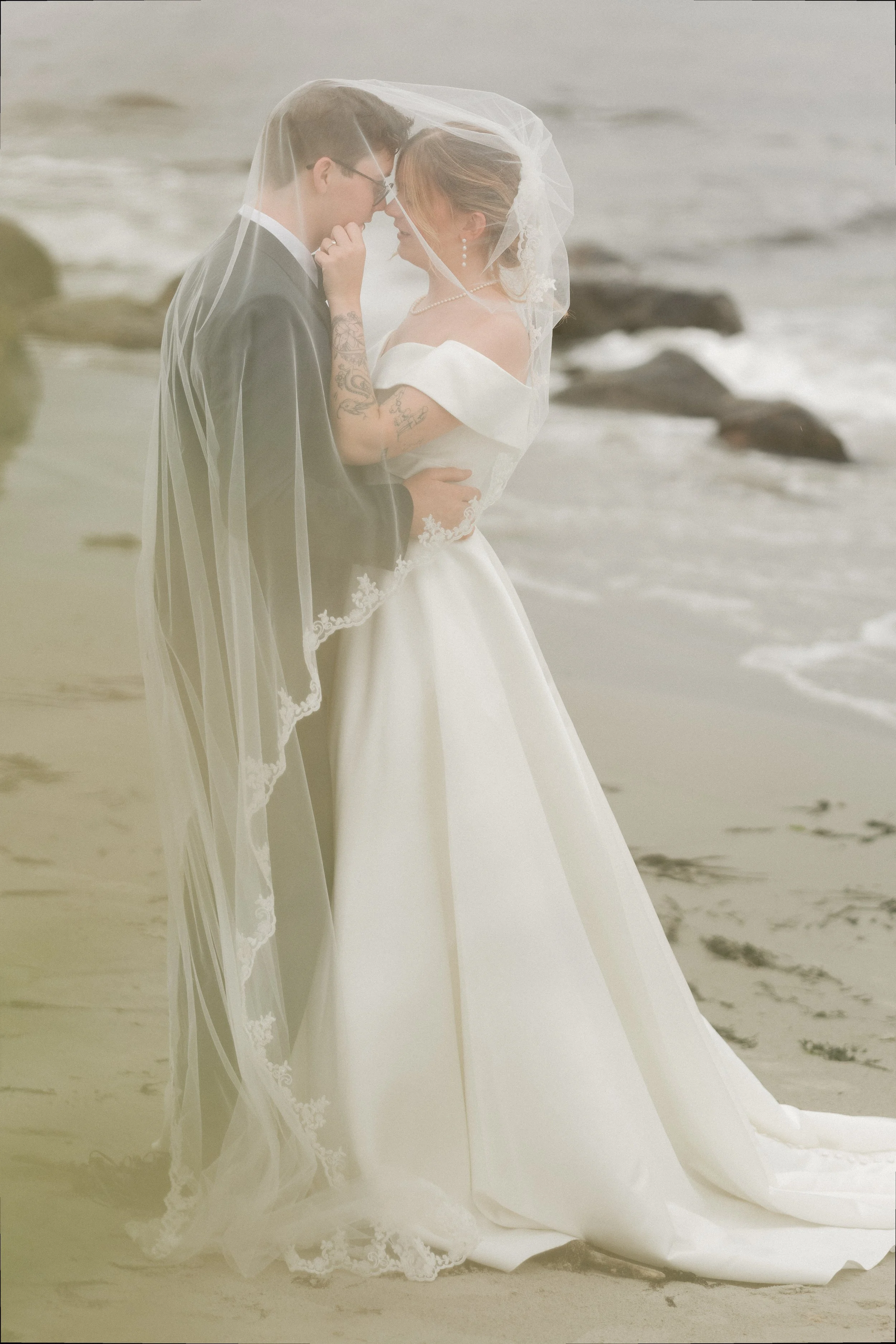 To personer i brudekjole og costume kysser på stranden, med vann og steiner i bakgrunnen.