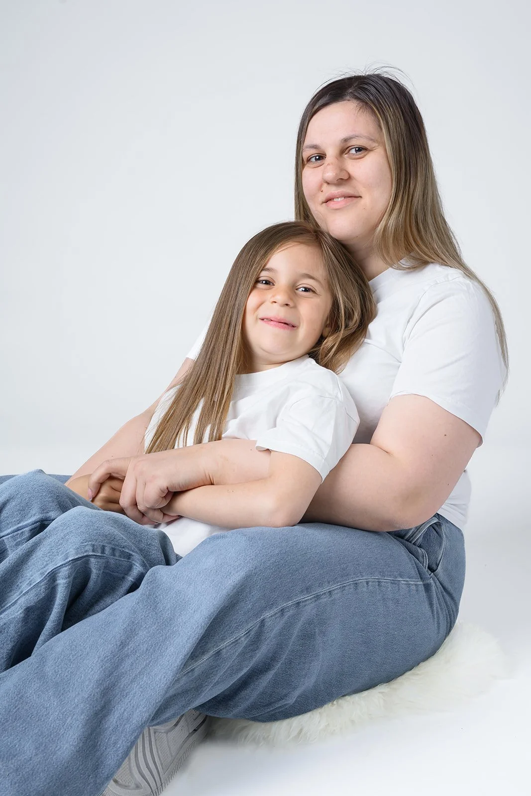 Mor og datter med langt brunt hår sitter smilende i hvite T-skjorter og blå jeans mot en lys bakgrunn.