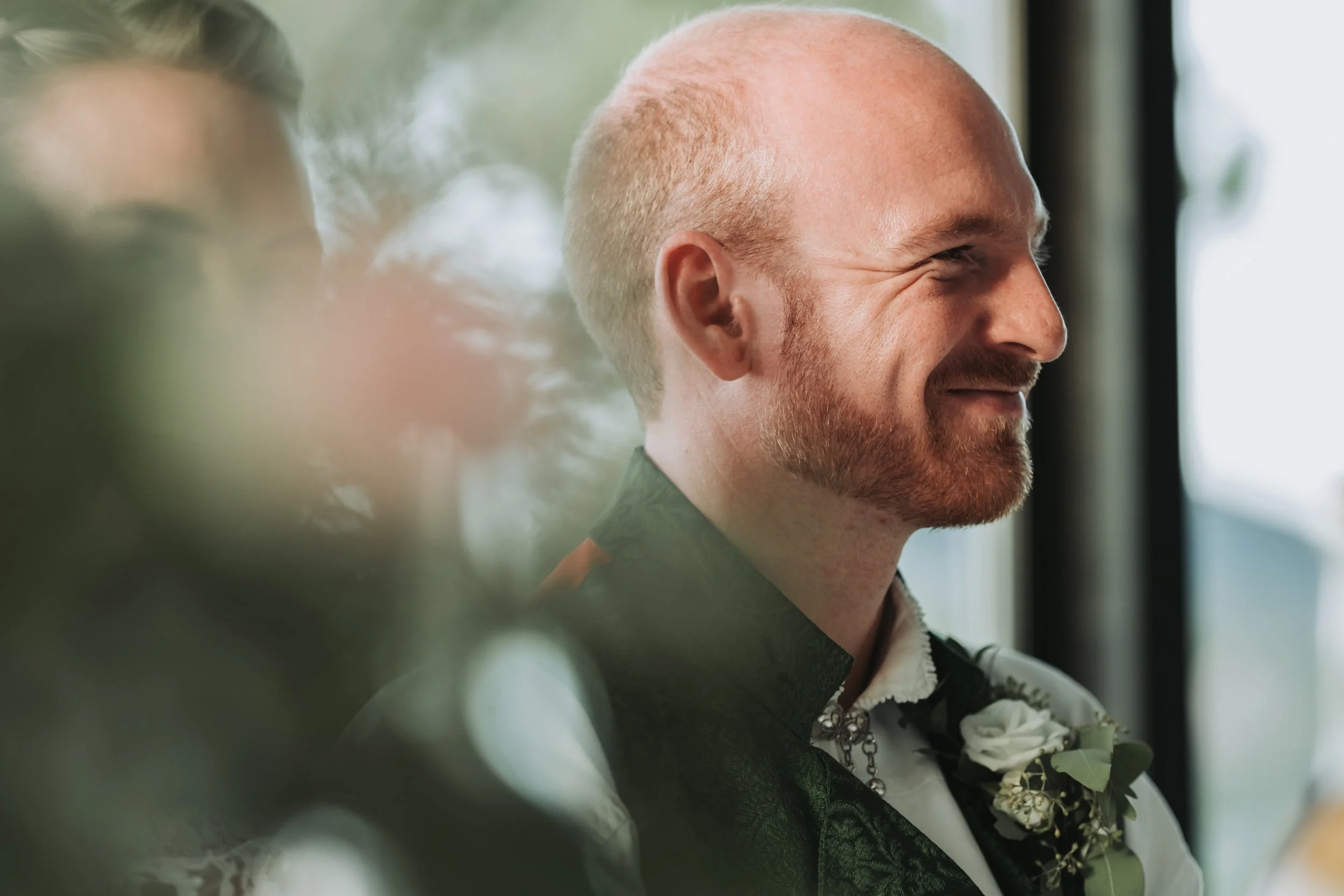 En mann med barbert hode som smiler når han sitter ved et vindu, iført ei svart vest og en hvit skjorte med en blomst som pynt.