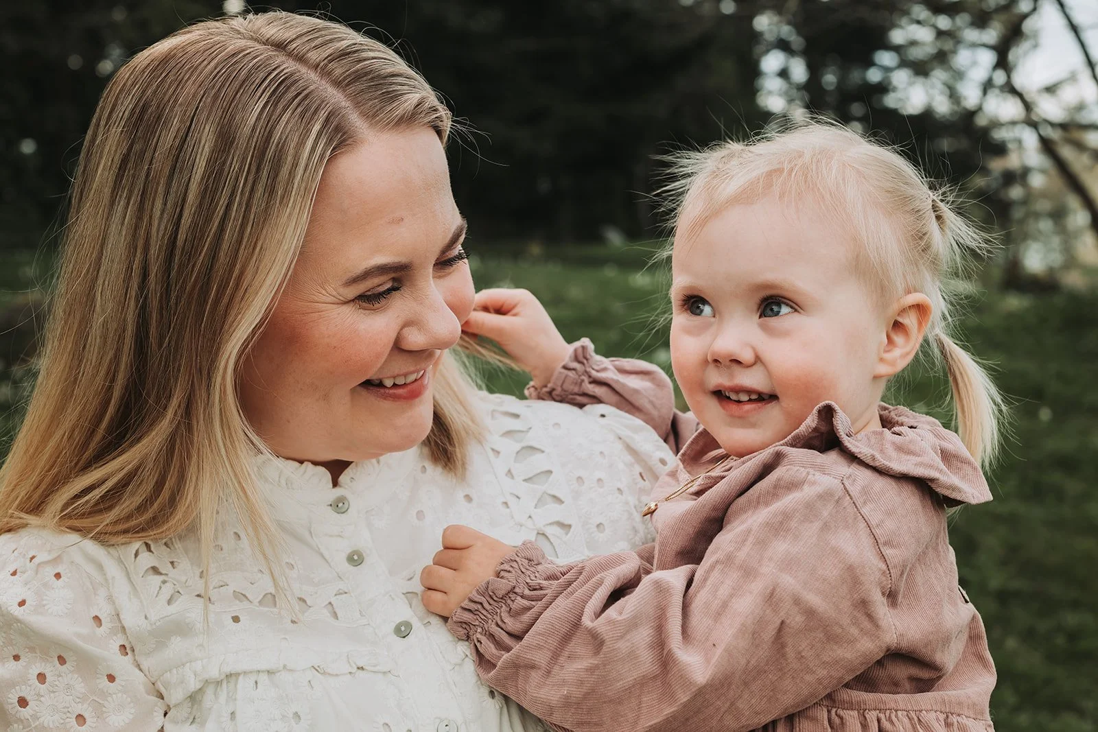 Smilende mor holder en liten datter utendørs i en grønn park.