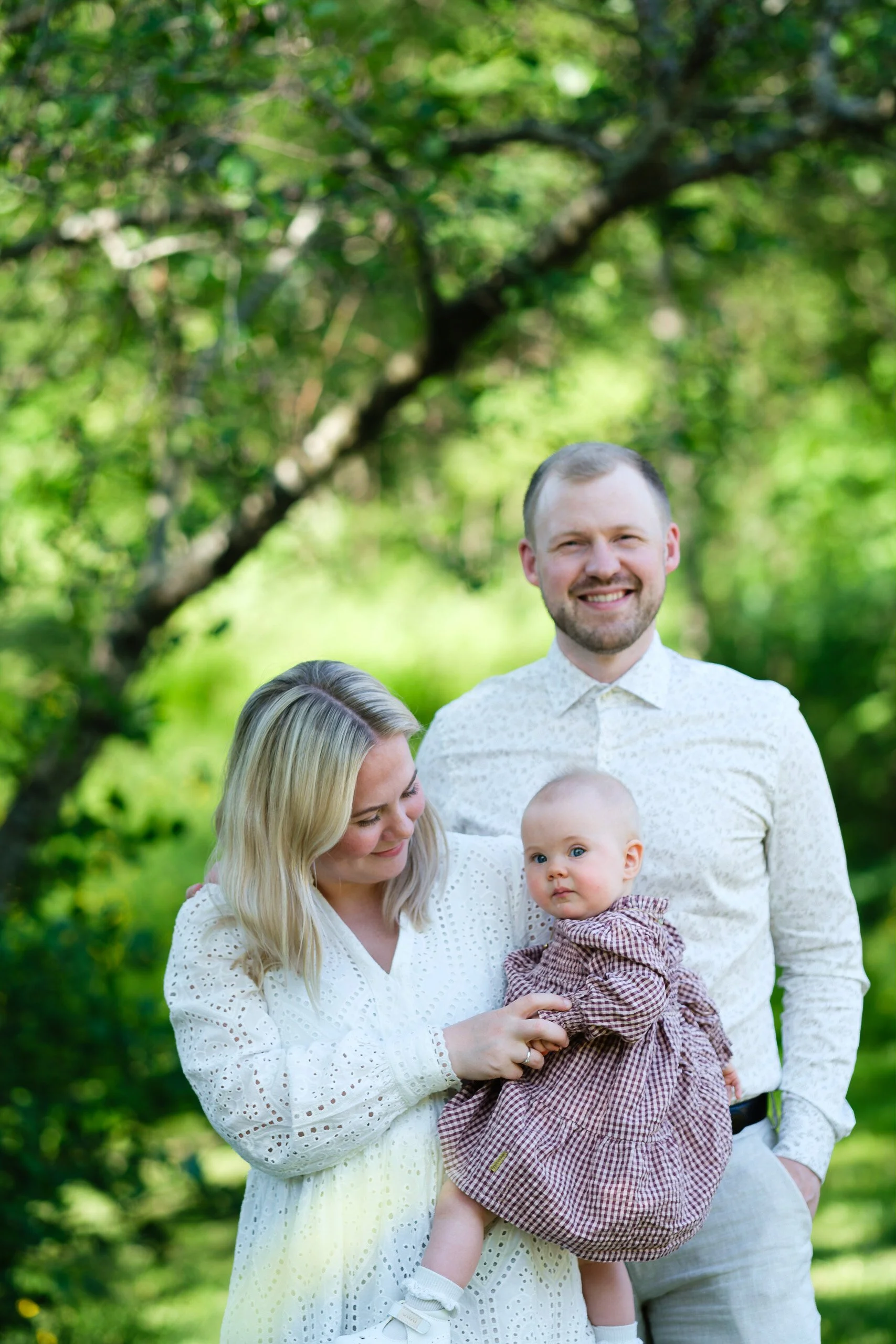 en familie av tre som står i en grønn park, smiler og ser mot kamera, barnet holdes av moren, en kvinne med blond hår og hvit kjole, mannen står bak dem i en lys skjorte. Bladene i bakgrunnen skaper en naturlig og lystig stemning.