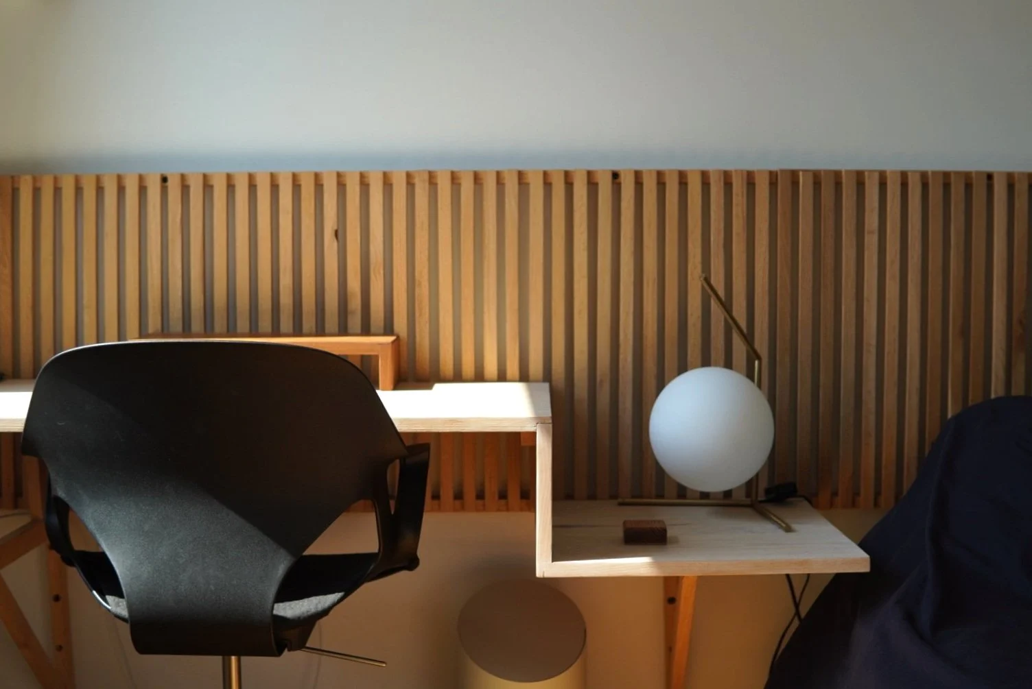 A minimalist workspace with a custom wooden desk, a black chair, a modern lamp with a round white shade, and a wooden slatted headboard or wall backdrop.