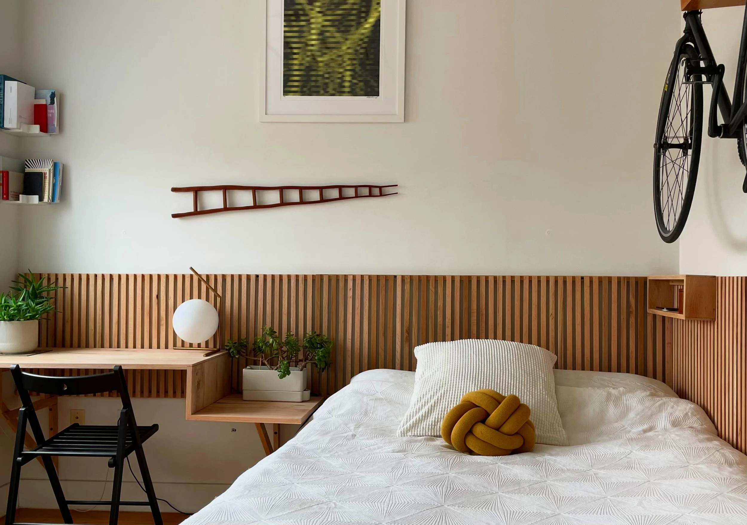 A bedroom with a wooden headboard, a bed with white bedding, and decorative pillows. To the left, there is a small desk with a potted plant and a black chair. Above the desk, a wall shelf holds books, and a small wooden ladder decoration is mounted o