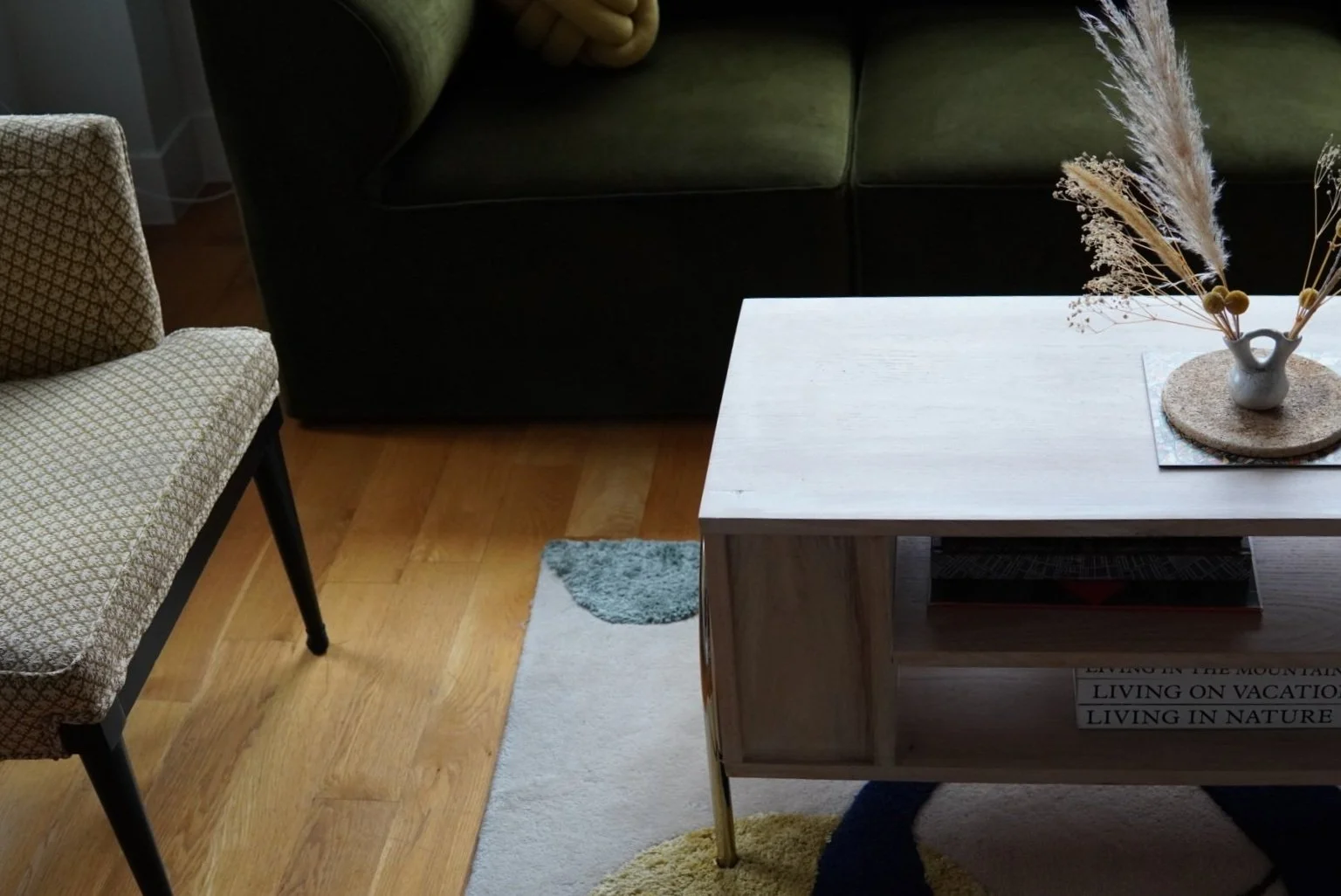 Living room with wooden floors, a green sofa, a patterned armchair, and a light-colored custom wooden coffee table with a vase of dried flowers on it. There are books on a shelf underneath the coffee table.