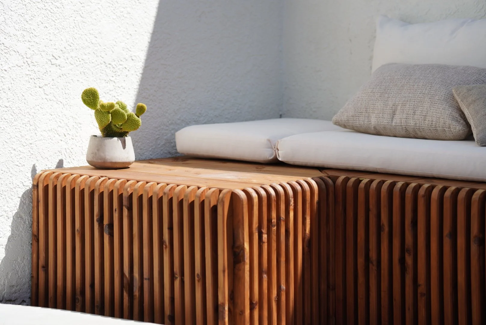 A custom wooden outdoor bench with white cushions and beige pillows next to a white stucco wall, with a small potted cactus plant on top.
