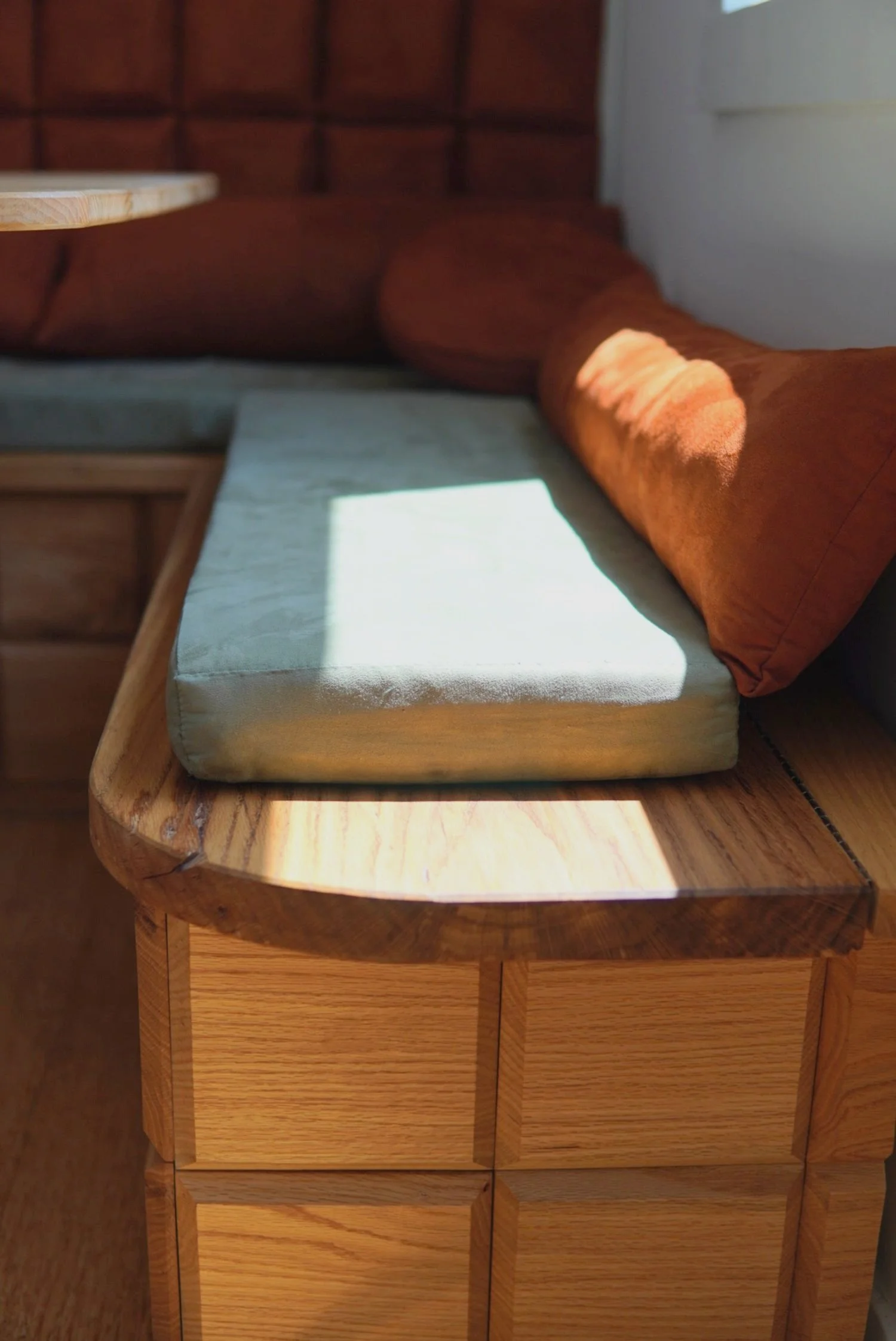 Close-up of a custom wooden storage bench with a light green cushion and an orange pillow, with sunlight casting shadows on the cushion.