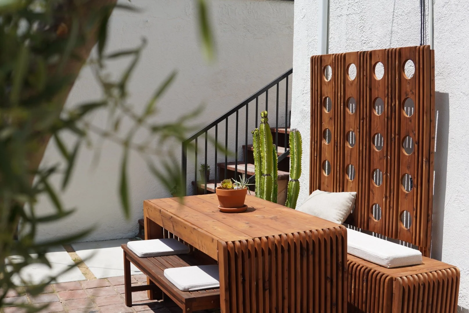 Custom outdoor patio table and bench with white cushions, a small plant in a terracotta pot, tall cacti, and a decorative wooden screen wall.