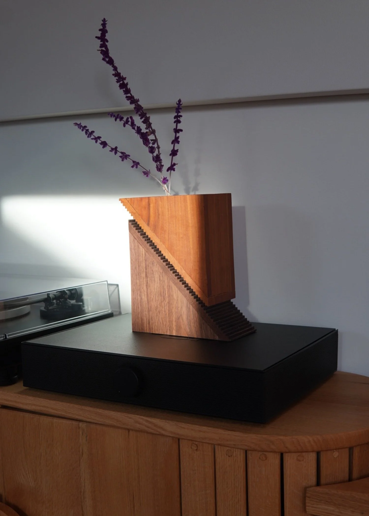 A wooden modern sculpture with a diagonal cut and small steps on one side, placed on a speaker on a wooden surface, with a purple flowering plant.
