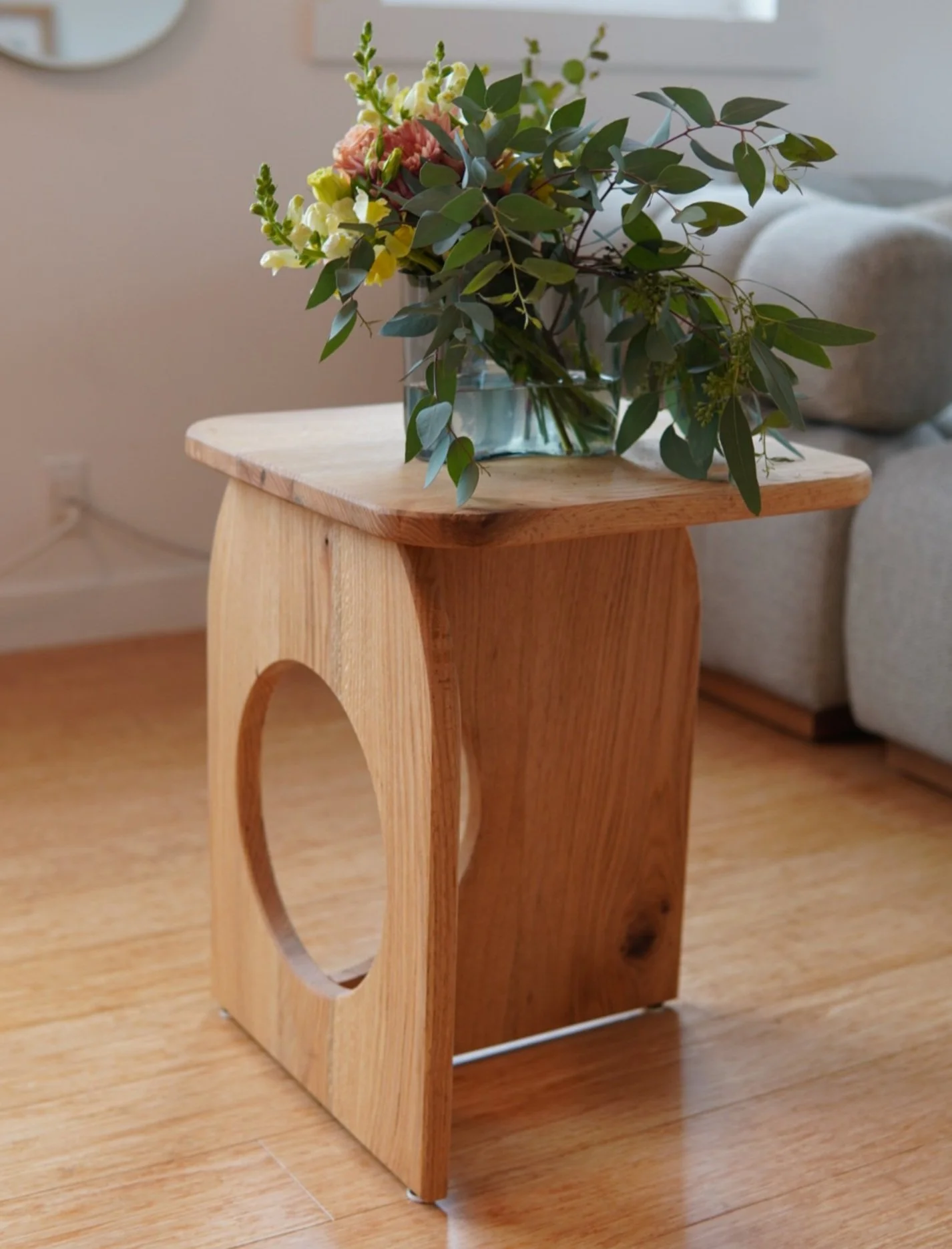 A custom wooden side table with a circular cutout design, holding a glass vase with a bouquet of pink, white, and yellow flowers along with green foliage, situated in a living room.