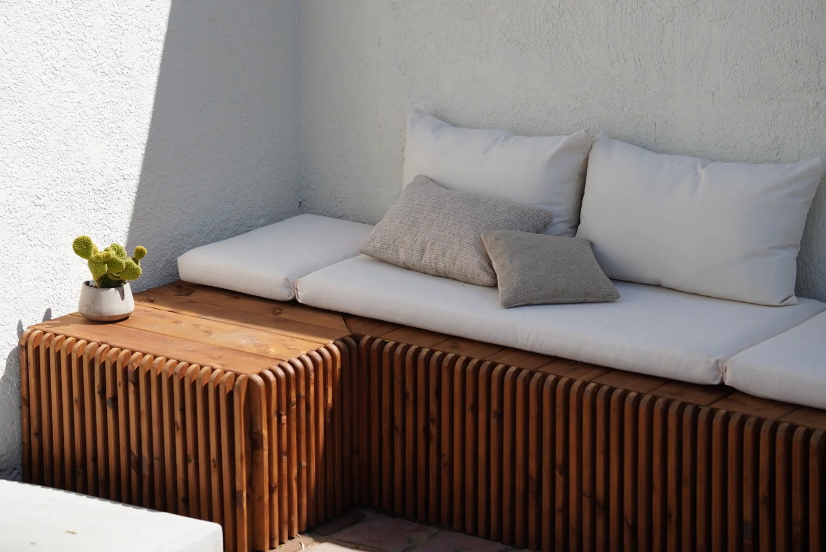 Custom outdoor wooden bench with beige cushions and pillows, accompanied by a small cactus plant in a white pot, against a white textured wall.
