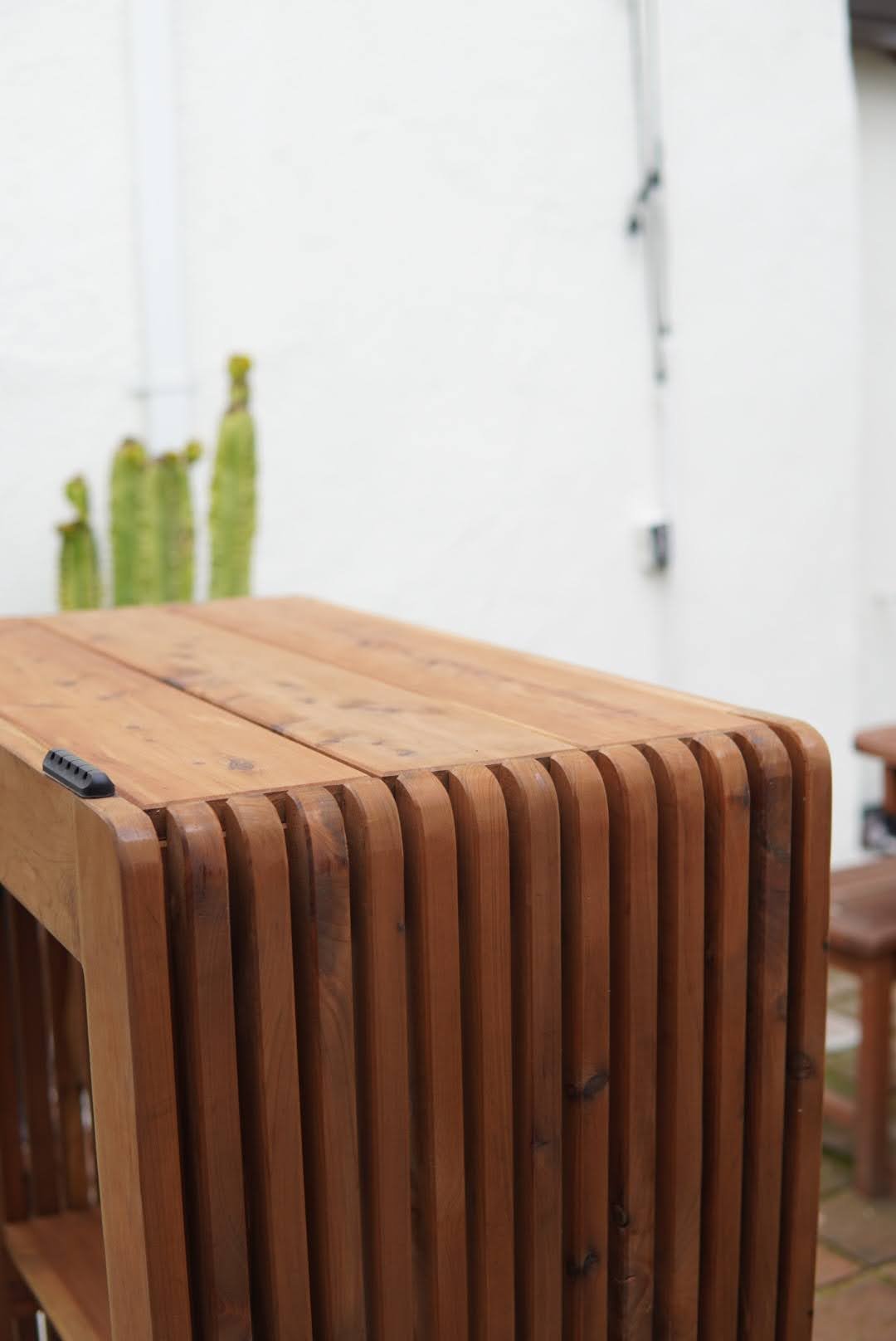 A custom wooden table with a slatted design is in the foreground, with a white wall and a small cactus plant in the background.