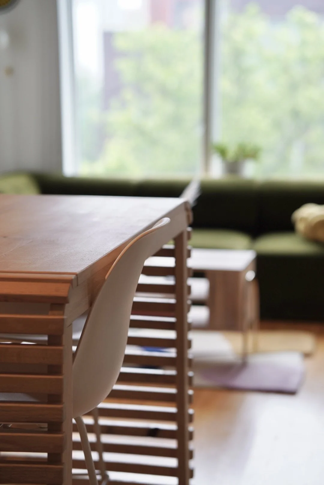 Close-up of a custom wooden dining table with a white chair in front, a window with greenery outside, a small table, and a sofa with pillows in the background.