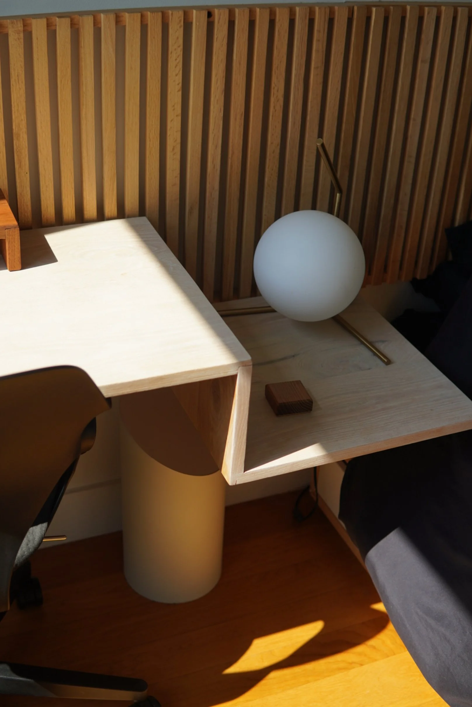 A custom oak desk with a black office chair. A spherical white lamp with a brass arm is on the desk, along with a small wooden block. The background features a custom wooden slat wall.