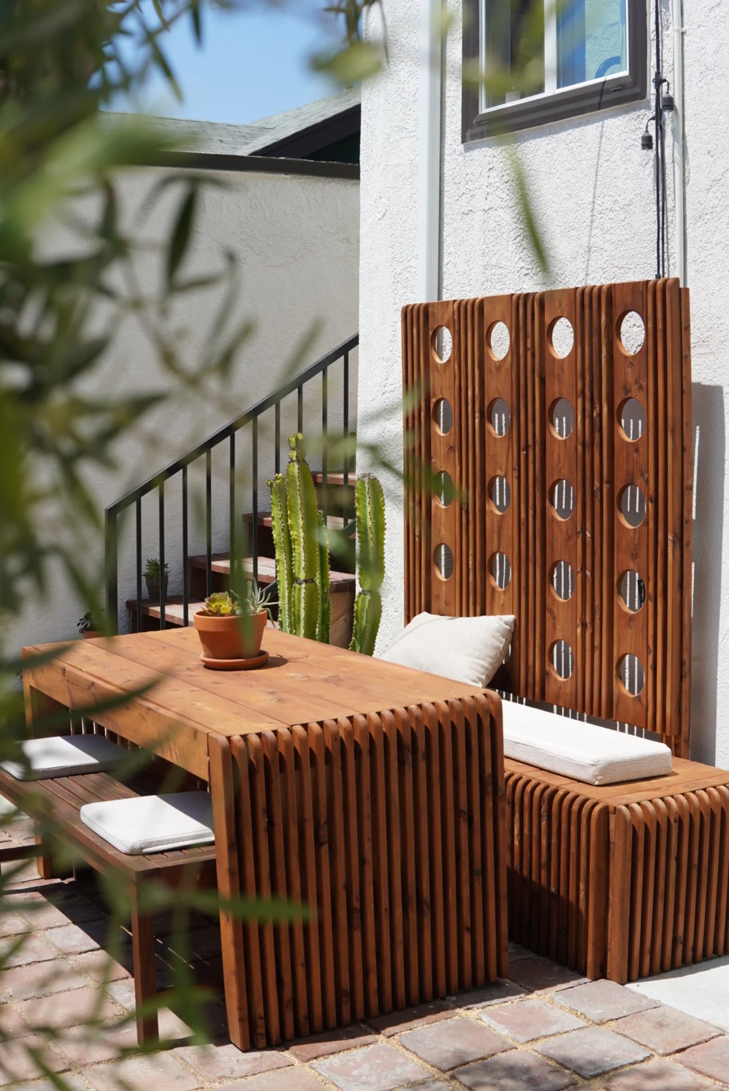 Outdoor patio seating area with custom wooden furniture, a cushioned bench, potted cacti, and a decorative wooden screen against a white wall.