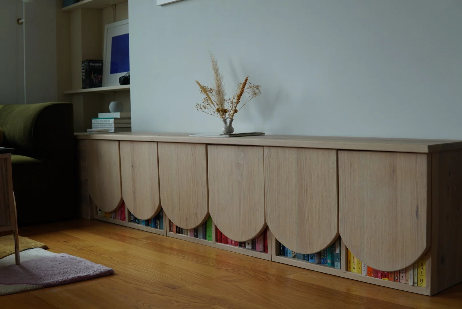 A modern custom wooden console with scalloped edges, filled with colorful books, topped with a small vase holding dried grasses, in a cozy living room.