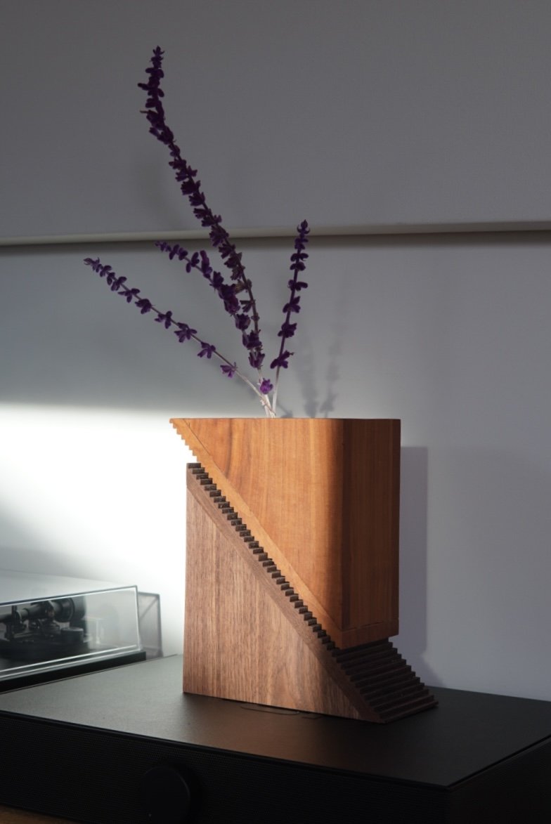 A modern wooden flower vase with purple flowers on top, placed on a dark surface against a gray wall.