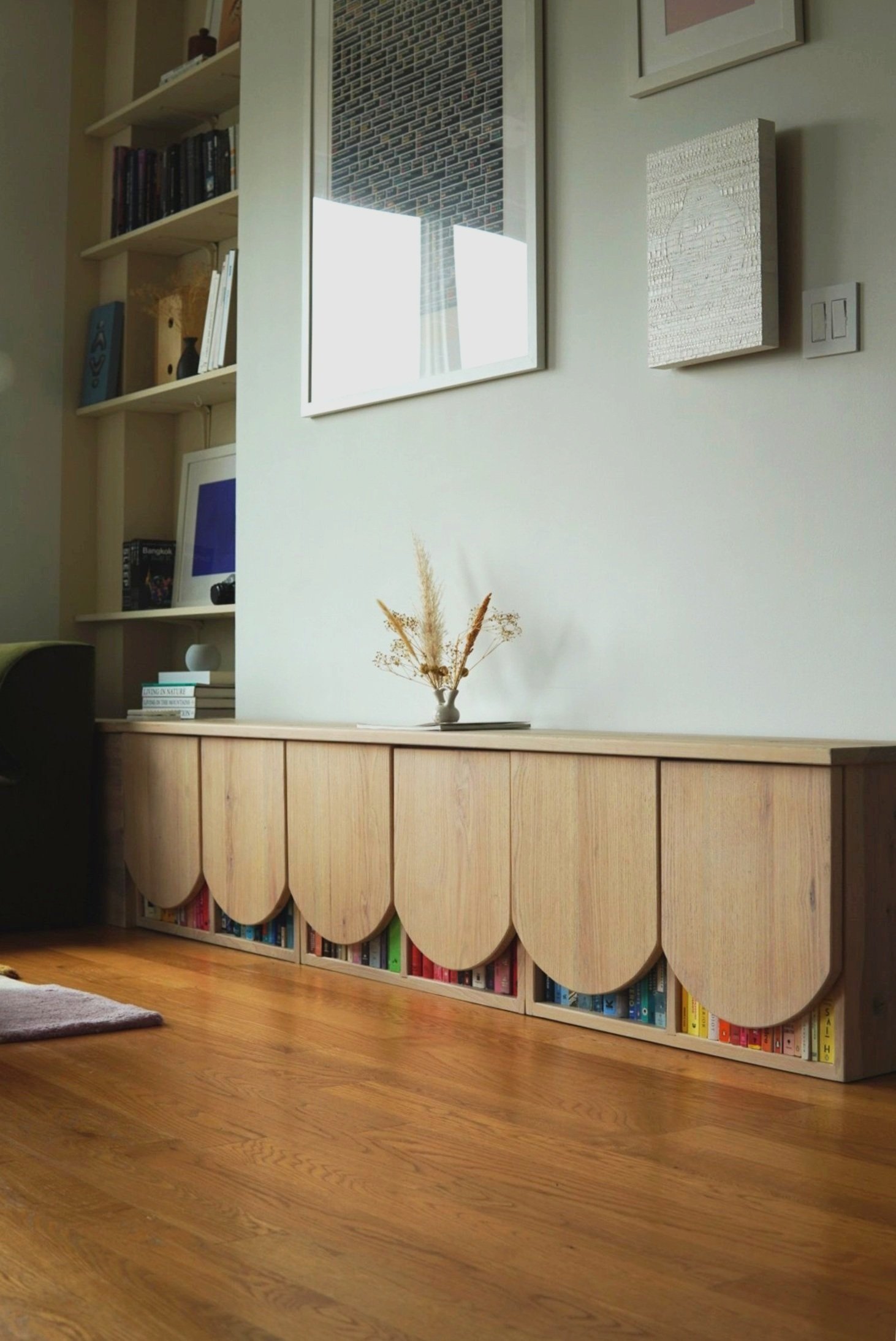 A custom wooden storage cabinet with a scalloped design at the bottom, featuring books with colorful spines underneath, in a living room with a hardwood floor, decorative wall art, and a small vase with dried plants on top.