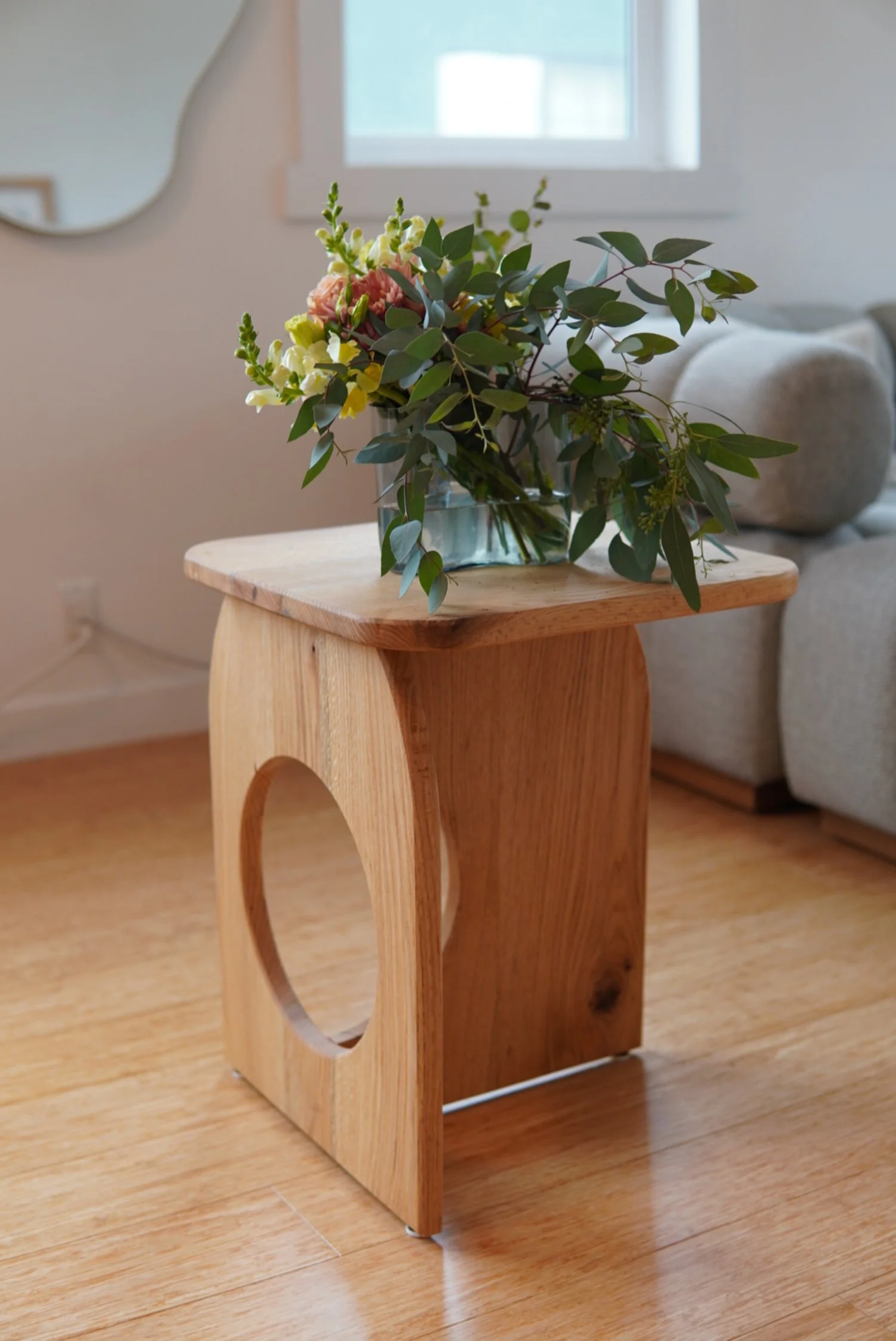Custom wooden side table with a circular cutout on the side, holding a glass vase filled with a bouquet of various green leaves and pink and white flowers in a cozy living room.