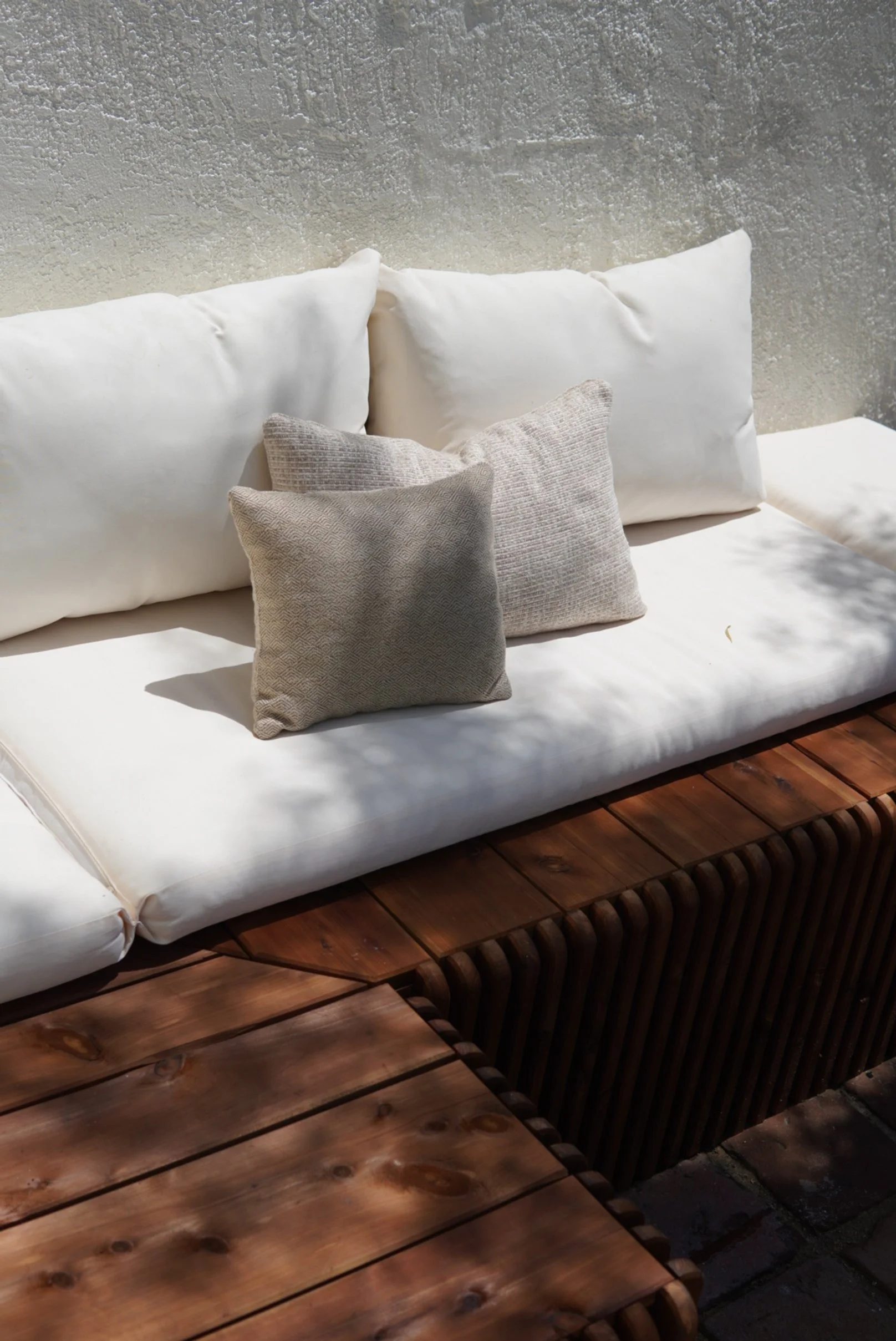 A white cushioned outdoor sofa with three beige textured pillows, situated on a wooden bench against a textured gray wall, with sunlight casting shadows.
