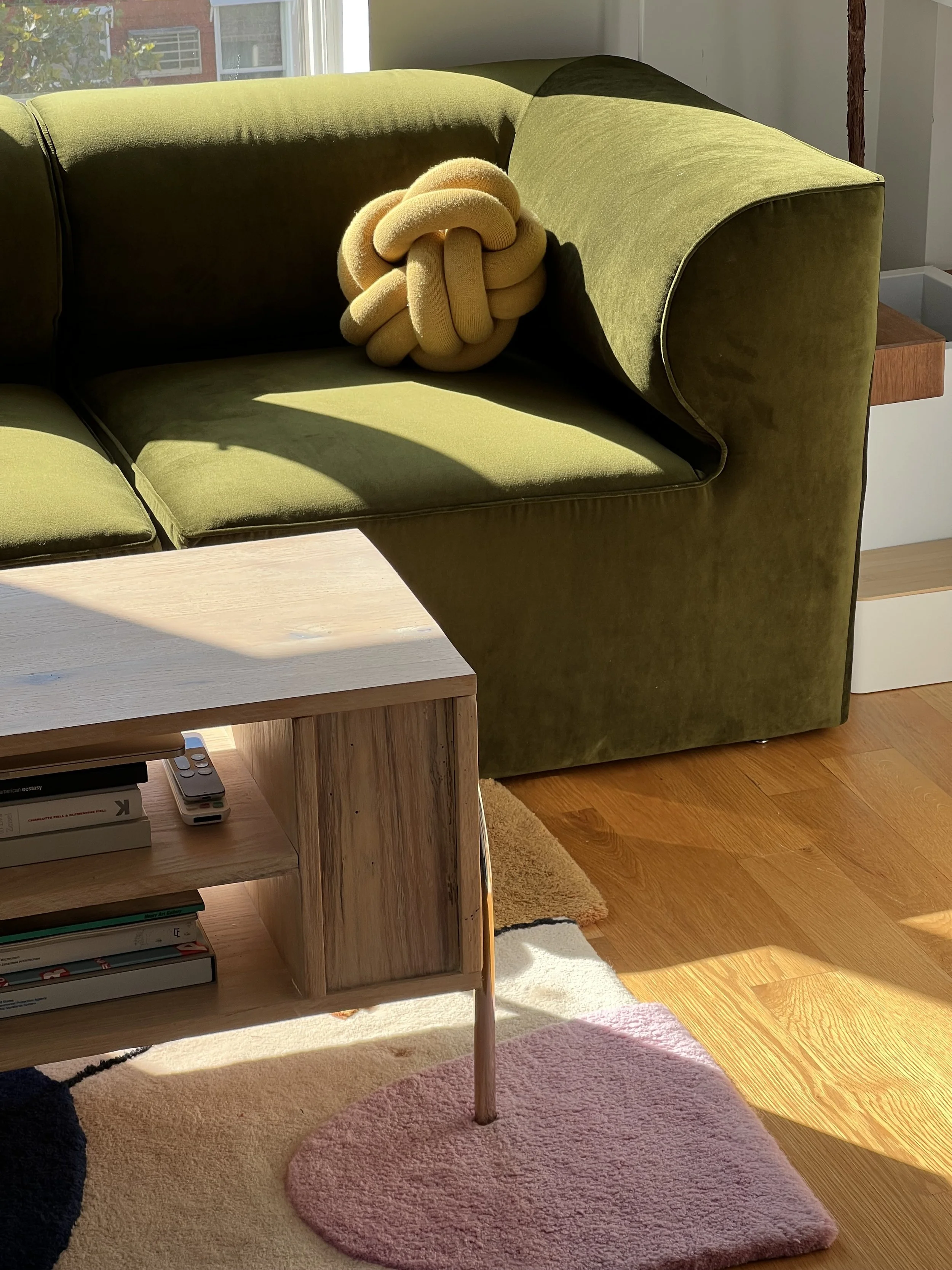 A green sofa with a yellow knot-shaped pillow on it, next to a custom wooden table, in a sunlit room with hardwood floors and a Proba rug.