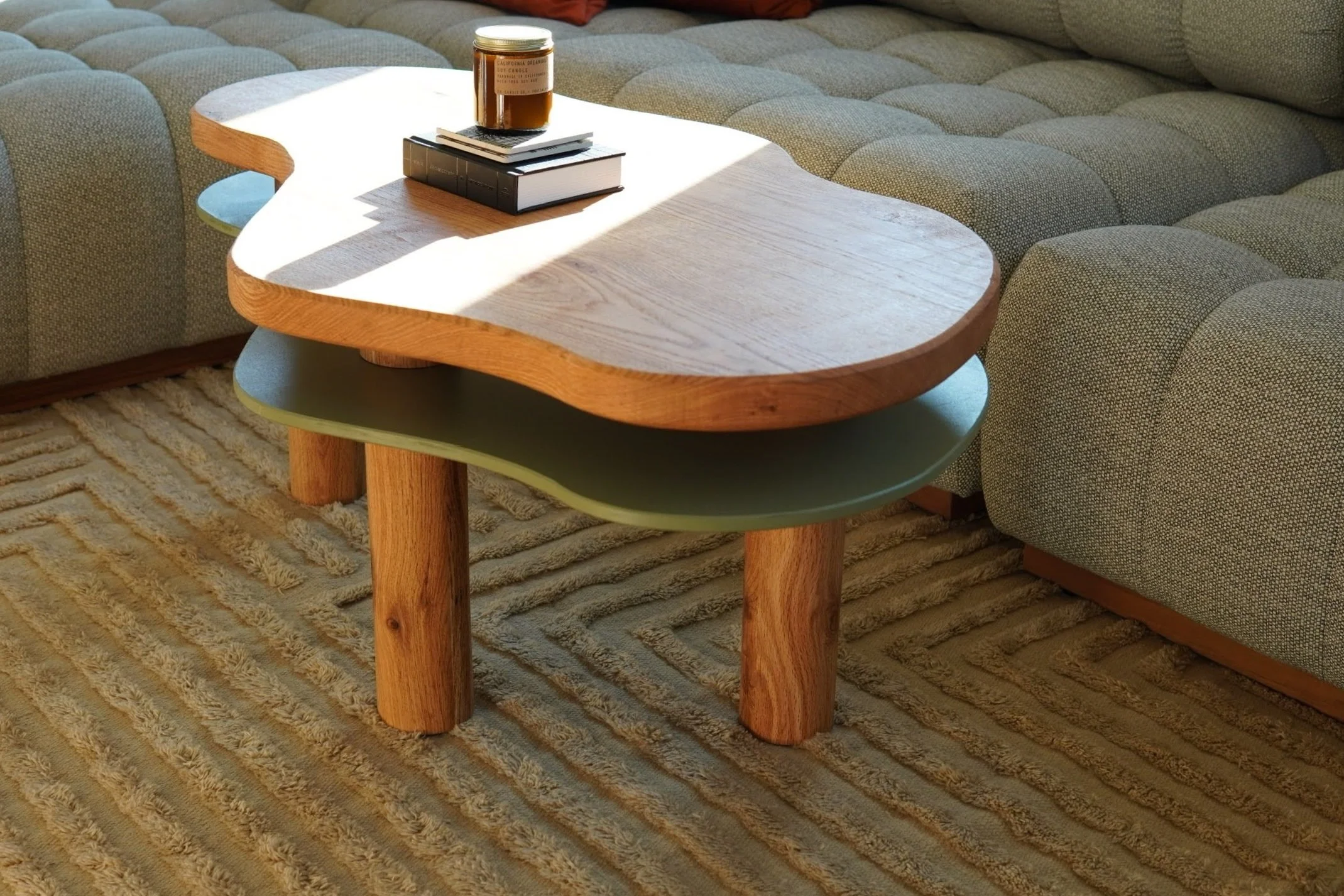 A living room with a modern wooden coffee table with rounded edges and an open shelf, placed on a beige patterned rug next to grey tufted sofas. The table has a jar, a couple of books, and a small stack of items on top.