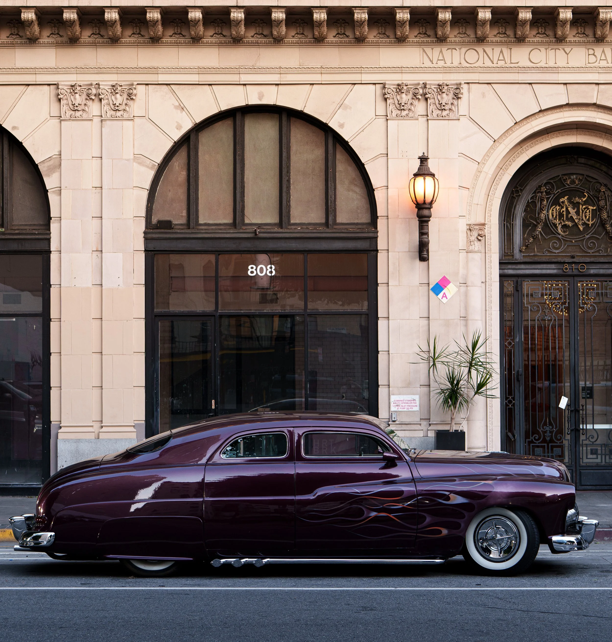 A highly modified purple 1951 Lincoln Mercury with blue and orange ghost flames in front of an art deco building