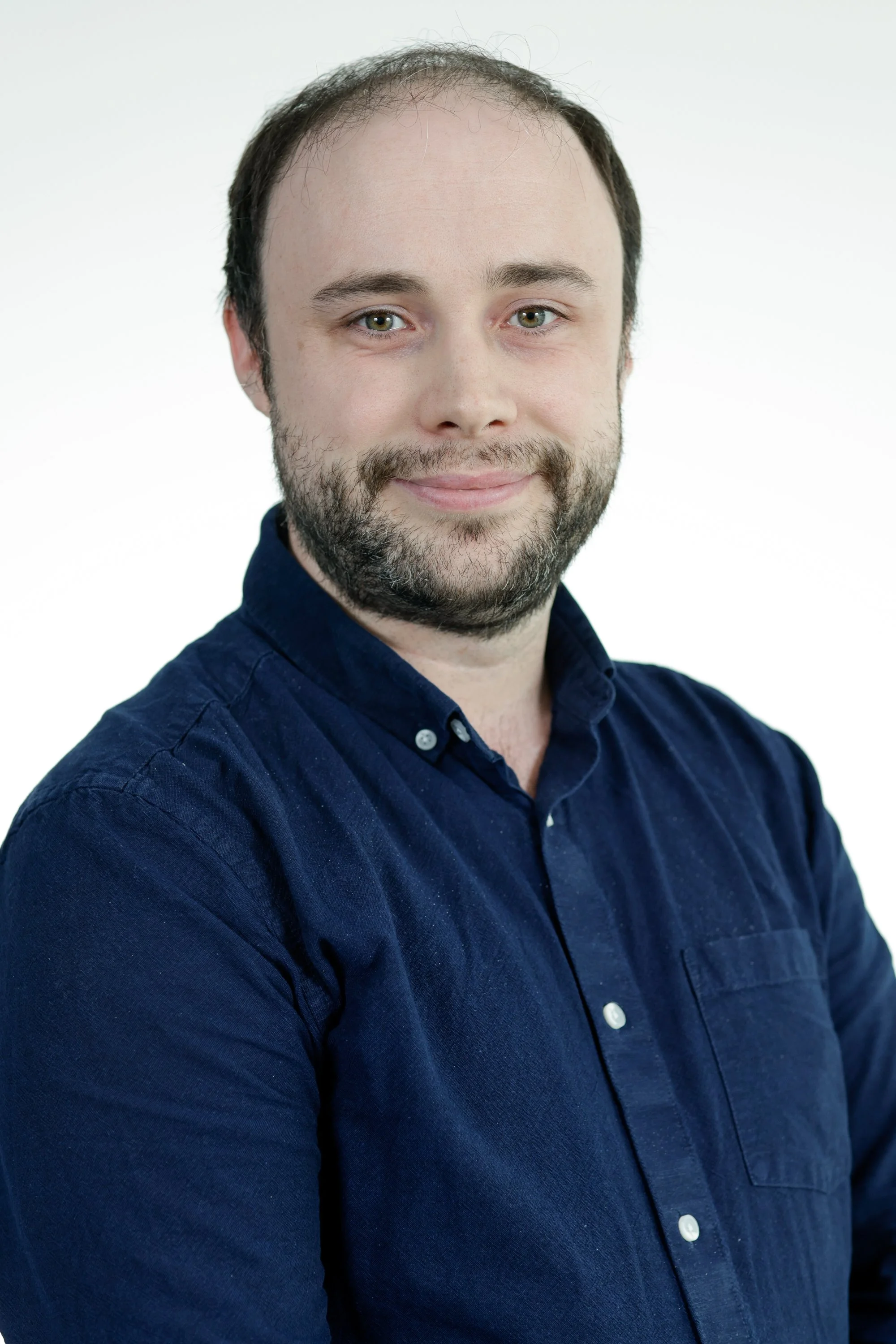 Headshot of a smiling young man with a beard and short dark hair, wearing a navy blue button-up shirt, posing against a plain white background.