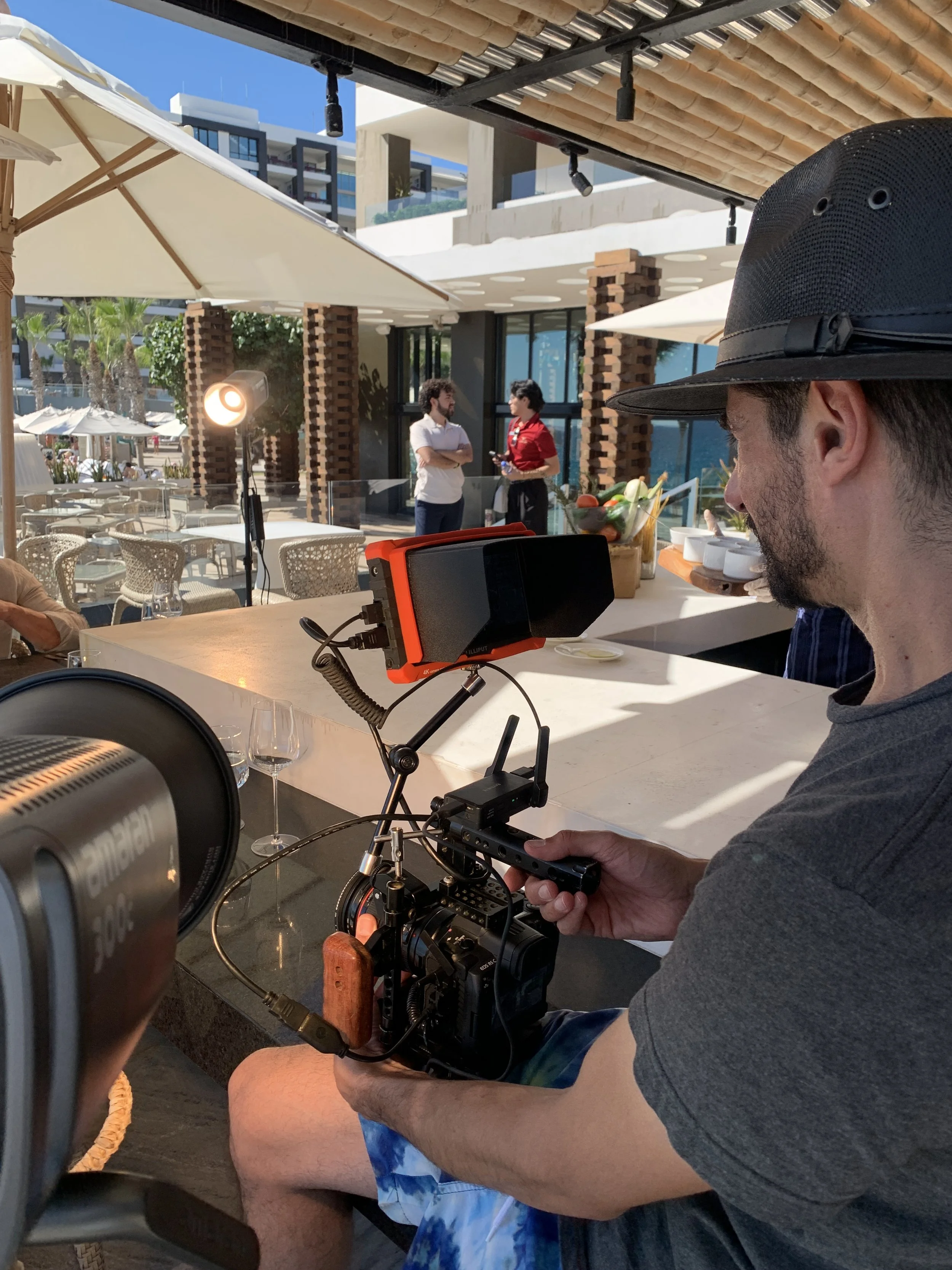 A man wearing a gray t-shirt and a black hat is filming with a professional camera at an outdoor restaurant or bar. In the background, two people are standing and talking near a counter, with outdoor furniture and umbrellas visible.