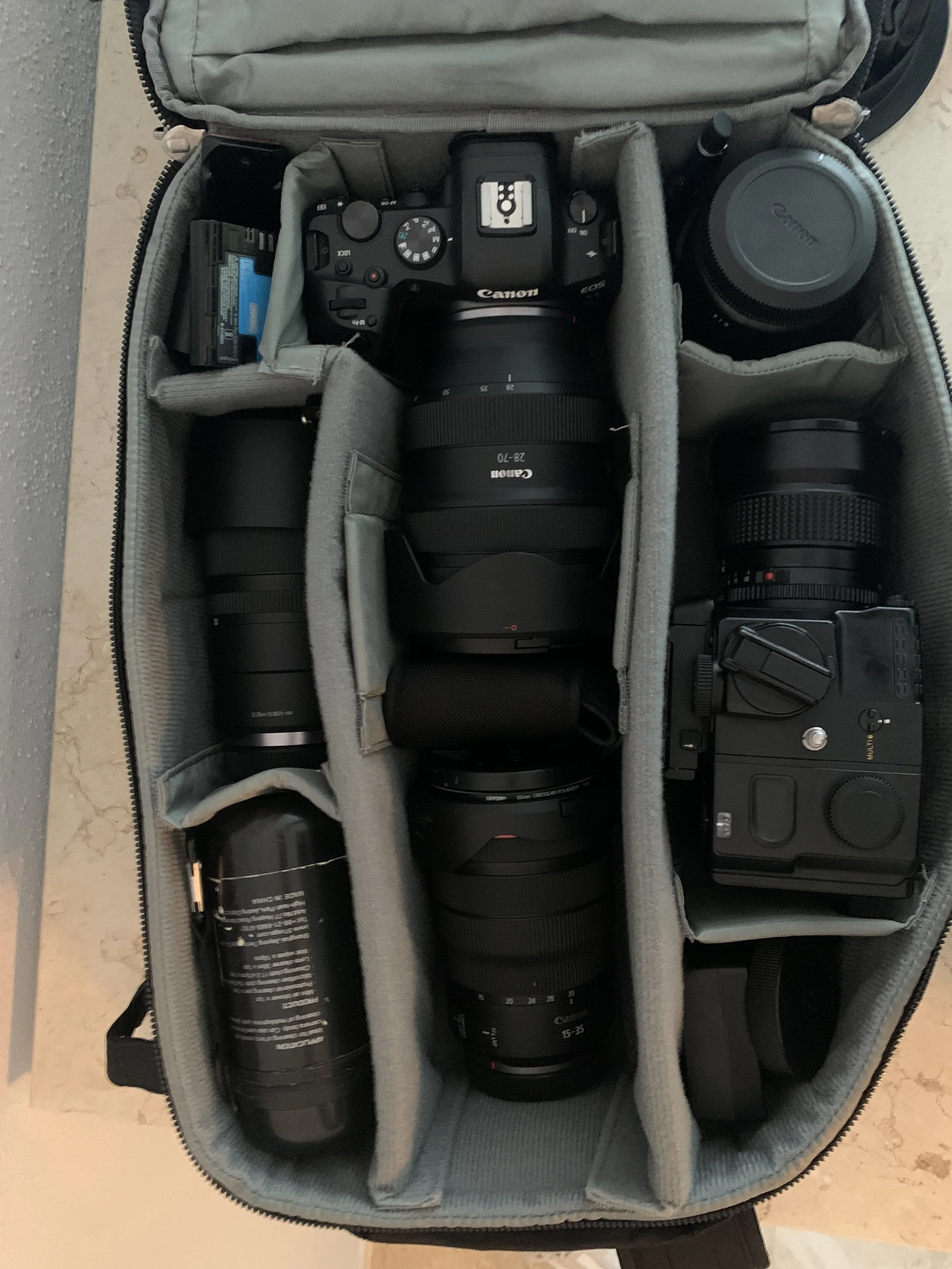 Open camera bag containing multiple camera lenses, a Canon camera with a lens attached, extra camera lenses, batteries, and other photography accessories.