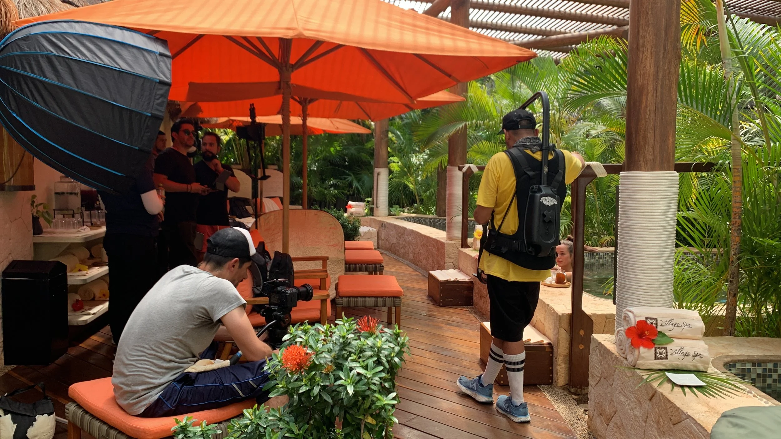 A behind-the-scenes look at a photo or video shoot in a tropical setting with orange umbrellas, crew members, and a woman in a pool.