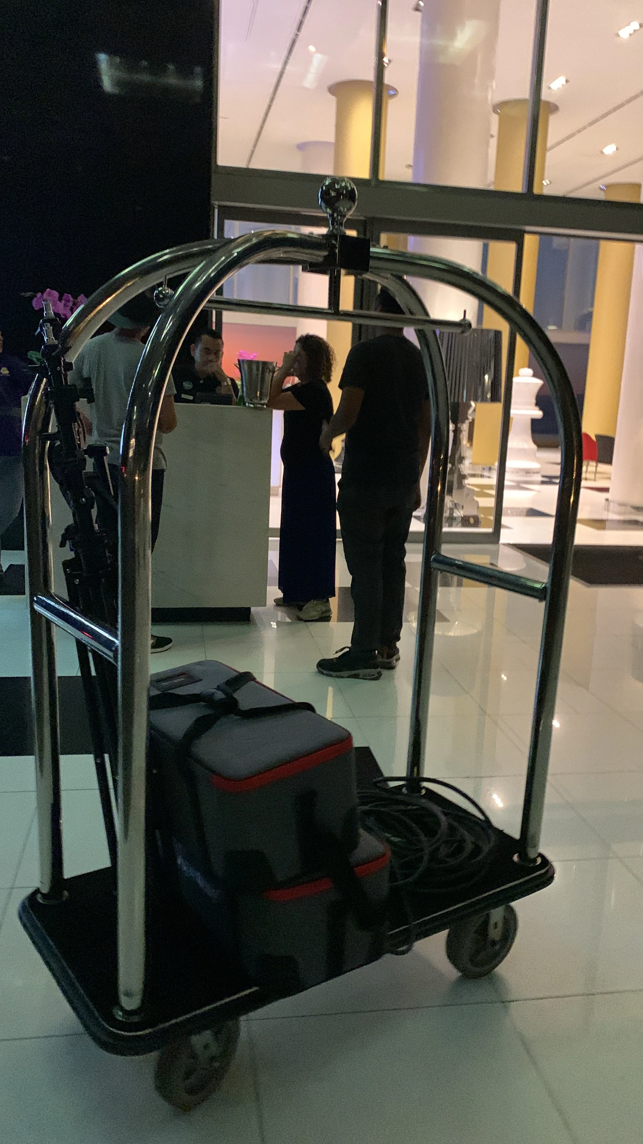 A hotel luggage cart with a black and red bag and coiled cables, positioned in the foreground, with a hotel reception desk and guests in the background.