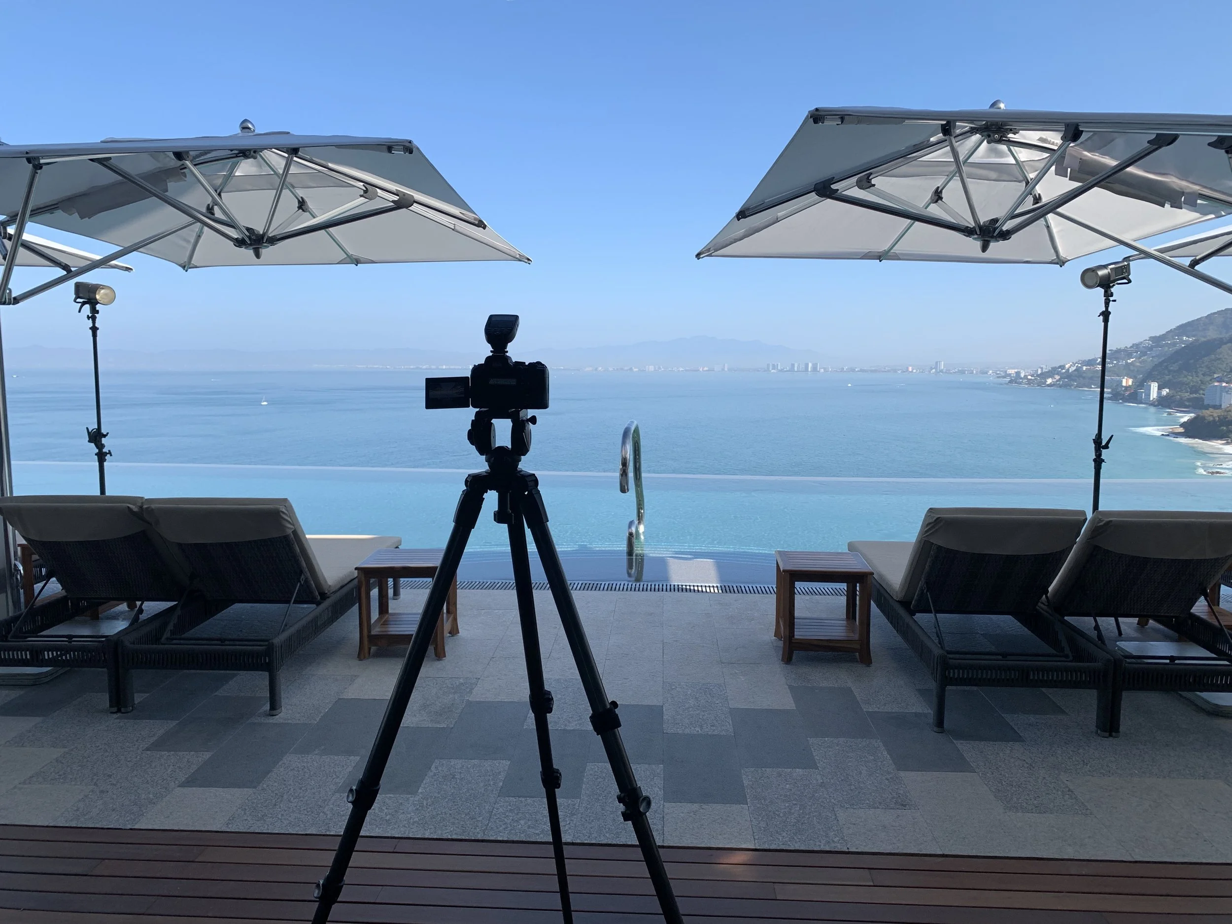 Balcony scene with two lounge chairs, two umbrellas, and a camera on a tripod, overlooking a large body of water with cityscape and mountains in the distance.