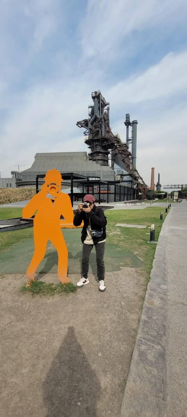 A woman taking a photo with a camera standing next to an orange cutout of a photographer silhouette at a museum or industrial site featuring a large, old industrial building with metal structures and smokestacks.