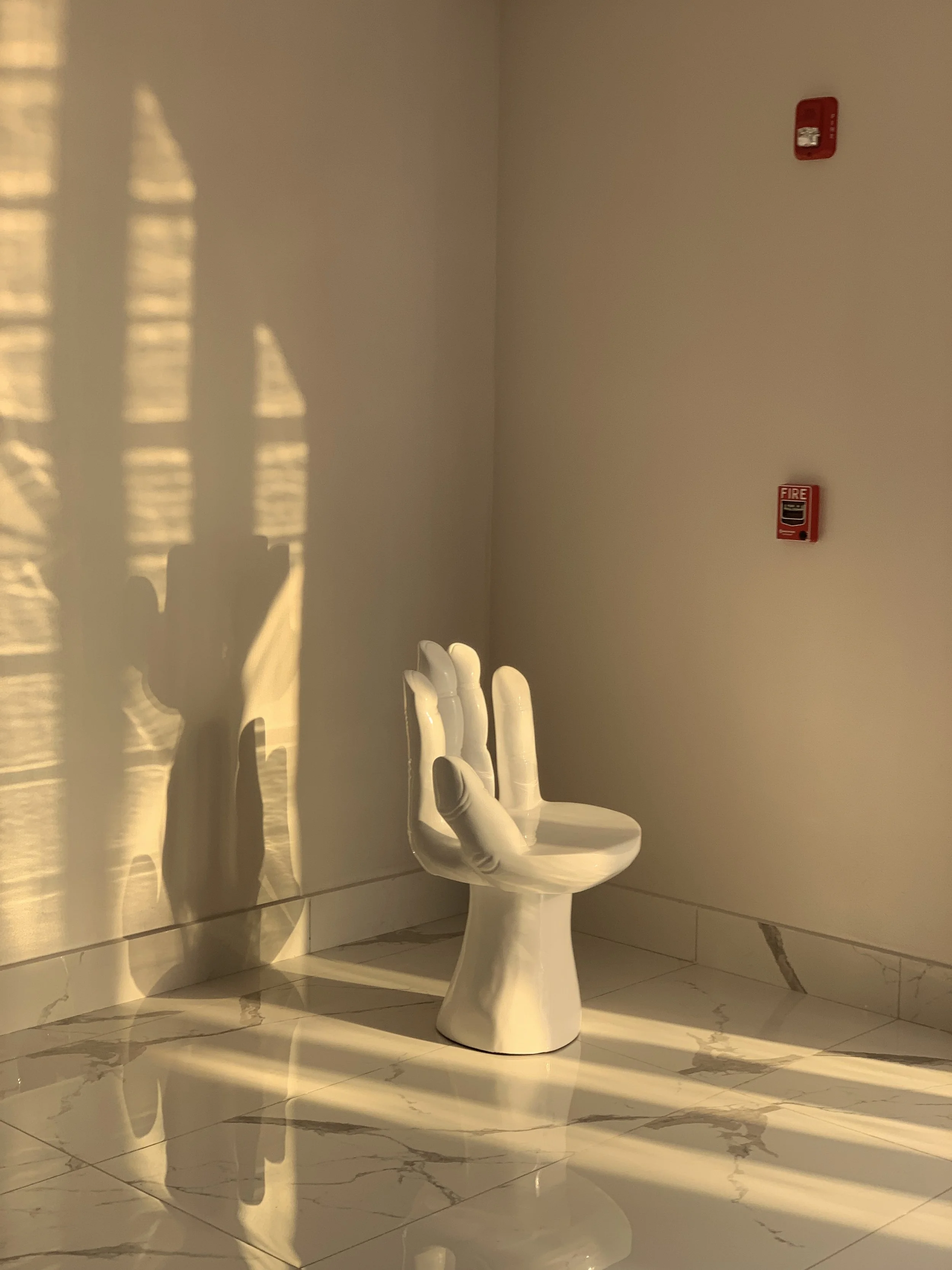 A white ceramic chair shaped like a hand with fingers as the backrest, positioned against a plain cream-colored wall. Shadows of window blinds cast on the wall and floor, with a fire alarm and smoke detector mounted on the wall.