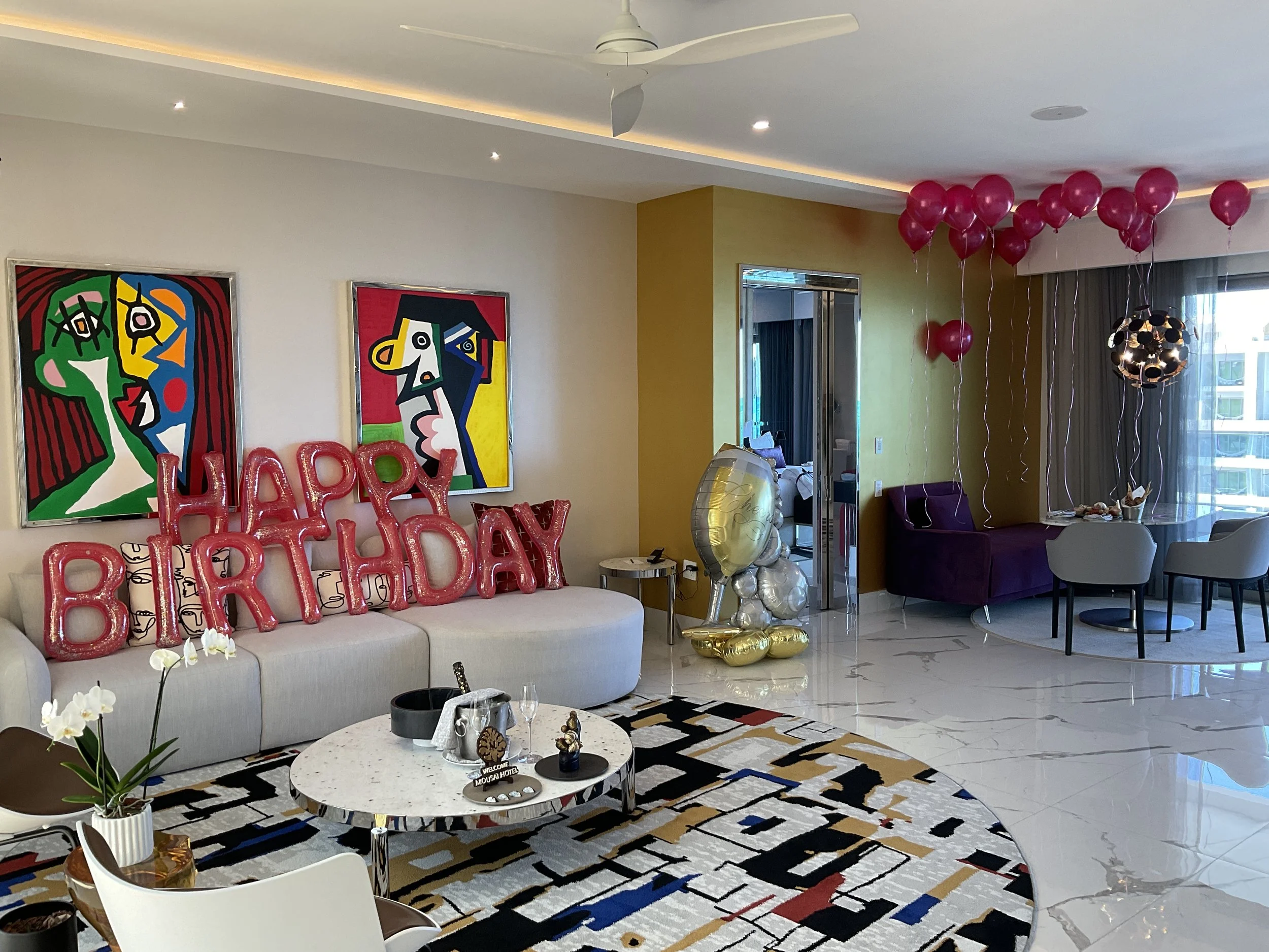 Living room decorated for birthday celebration with pink balloon letters spelling 'HAPPY BIRTHDAY', colorful balloon clusters, modern abstract paintings on the wall, a white sofa, a coffee table with floral decorations, and a multicolored patterned rug.