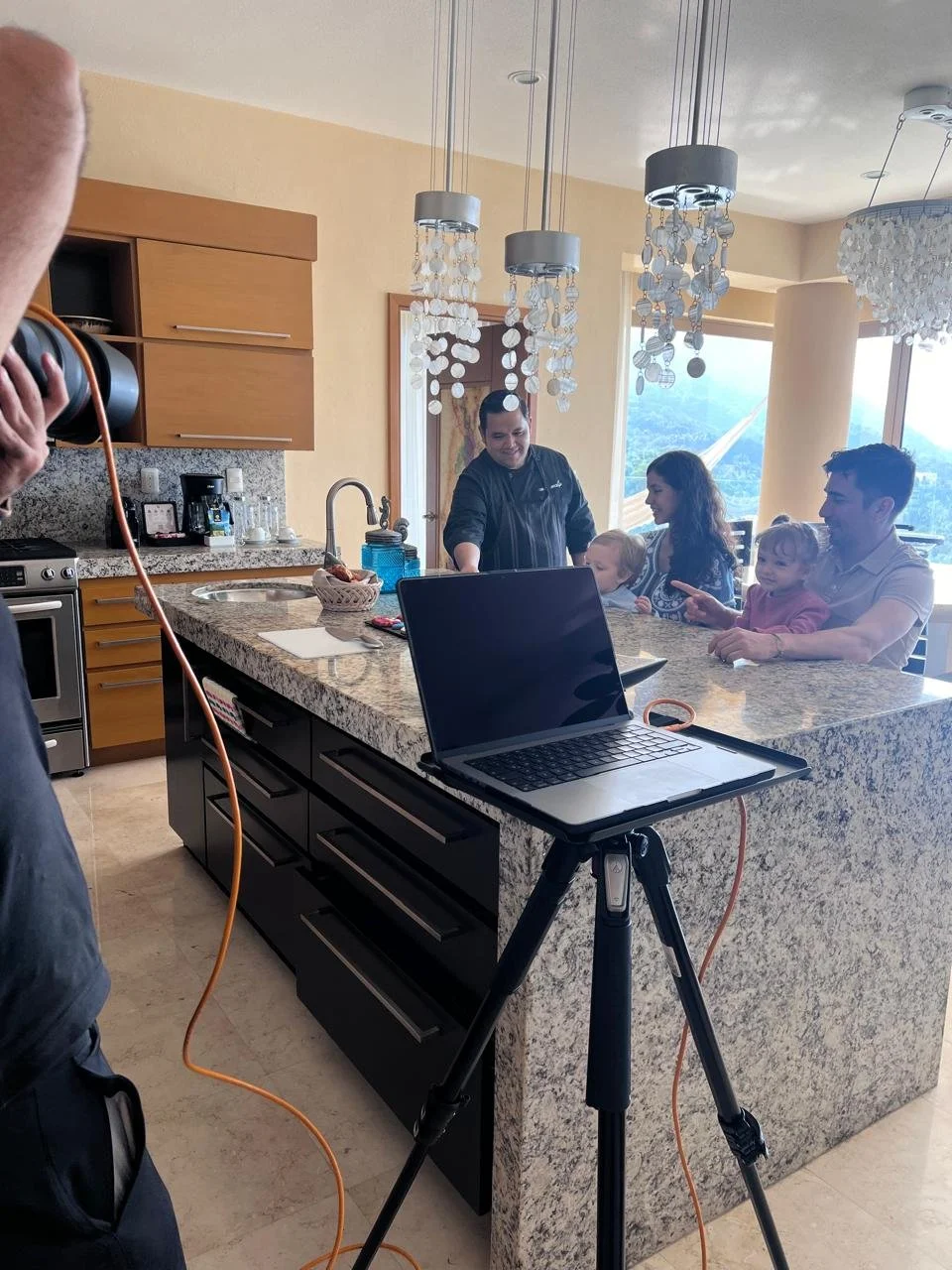 A family photo shoot happening in a modern kitchen with a granite island. Four people are sitting on the counter, including two children and two adults. A photographer is taking the picture, and there's a laptop on a tripod nearby. There are three hanging chandeliers overhead and a large window in the background showing a scenic view.