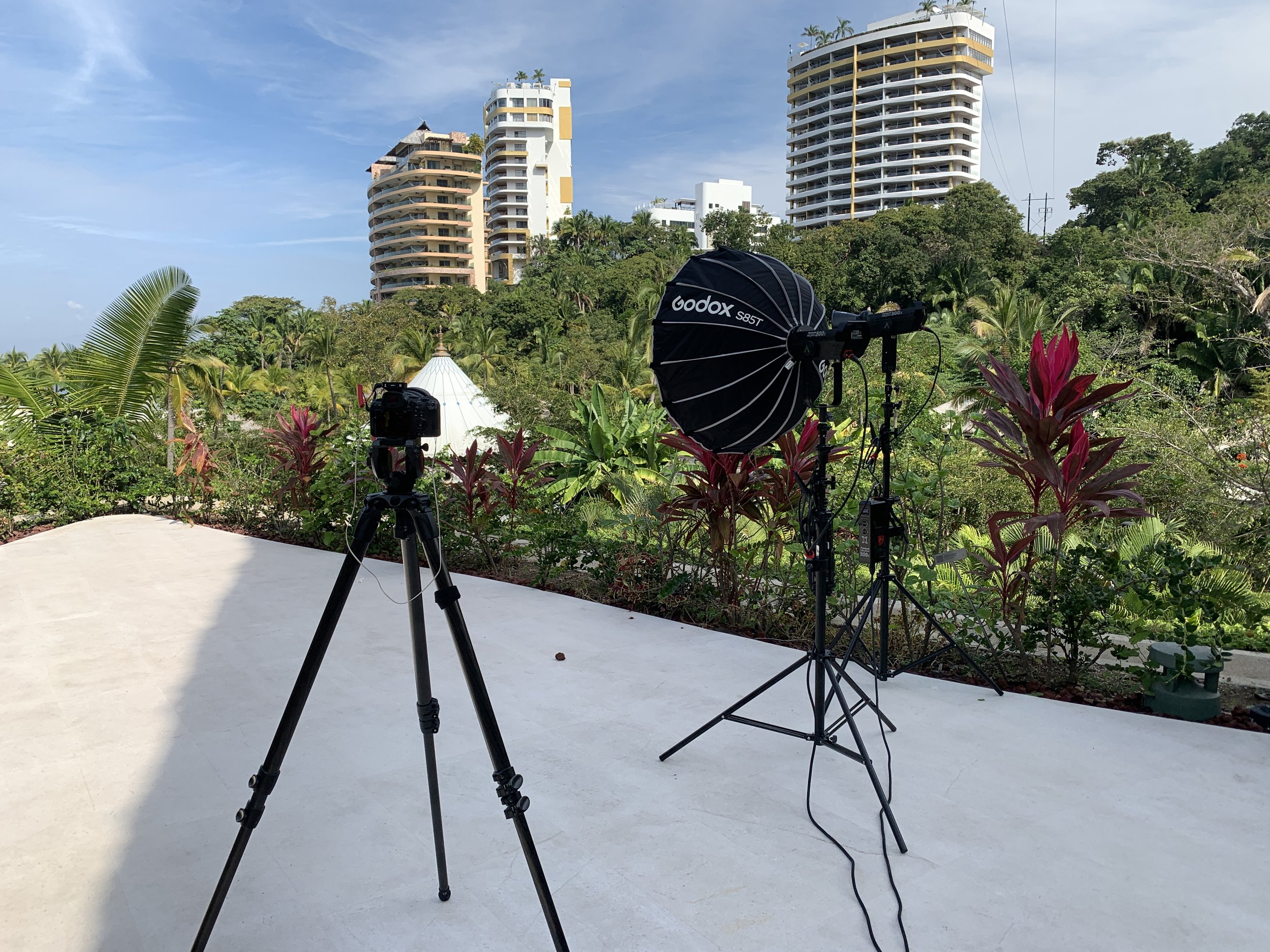 Photography setup with a camera on a tripod and a softbox light in an outdoor terrace with tropical plants, high-rise buildings, and a partly cloudy sky in the background.