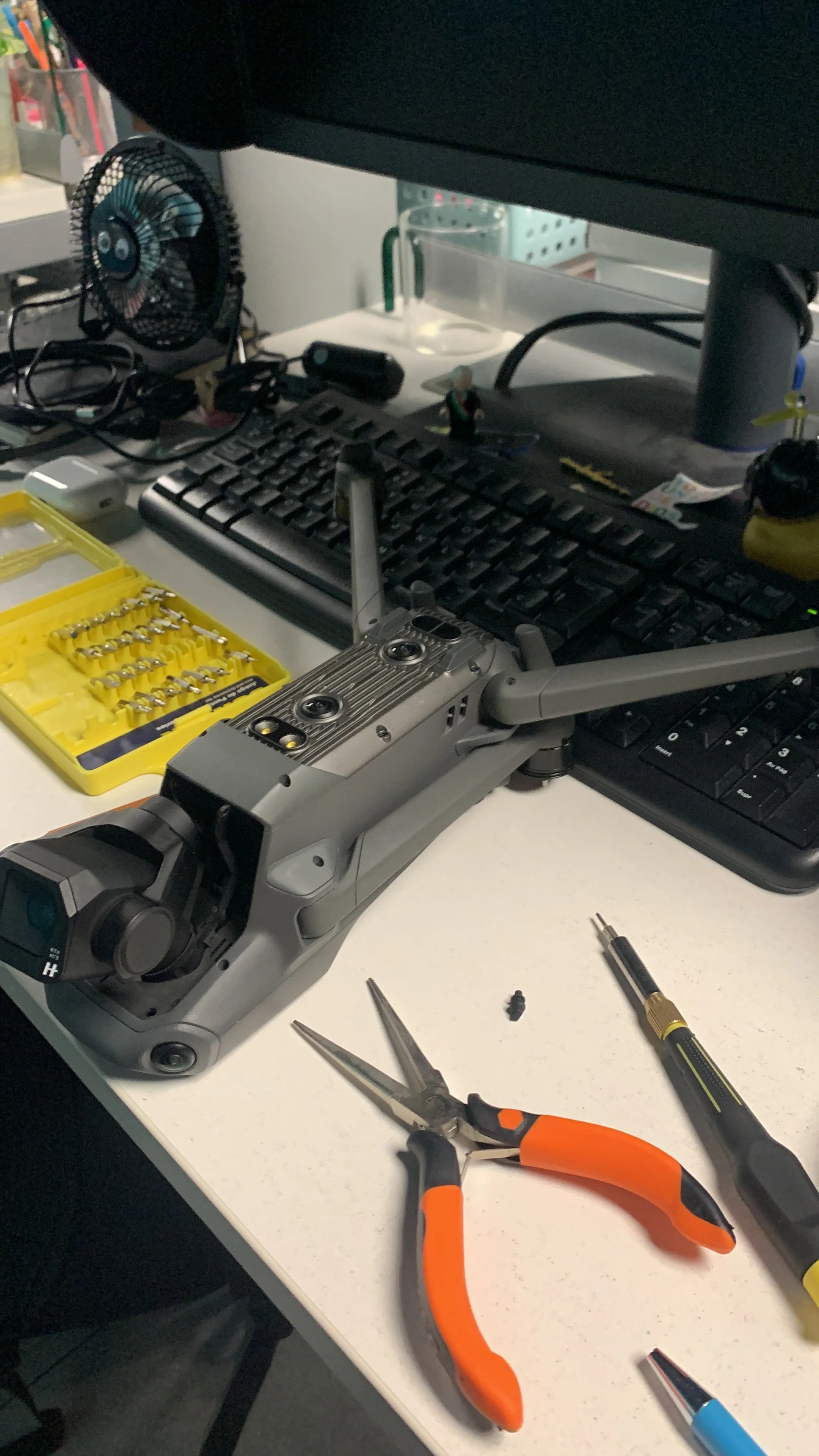 Workstation with a drone, tools, and electronic components on a white desk. There's a set of screwdriver bits, a small pair of pliers with orange handles, a screwdriver, a computer keyboard, and a monitor.