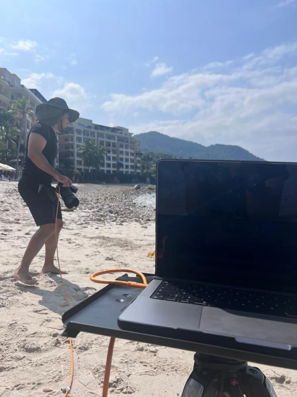 A woman wearing a sun hat and black clothing walking on the sandy beach, holding a camera, with buildings and mountains in the background. A laptop is set up on a tripod in the foreground.