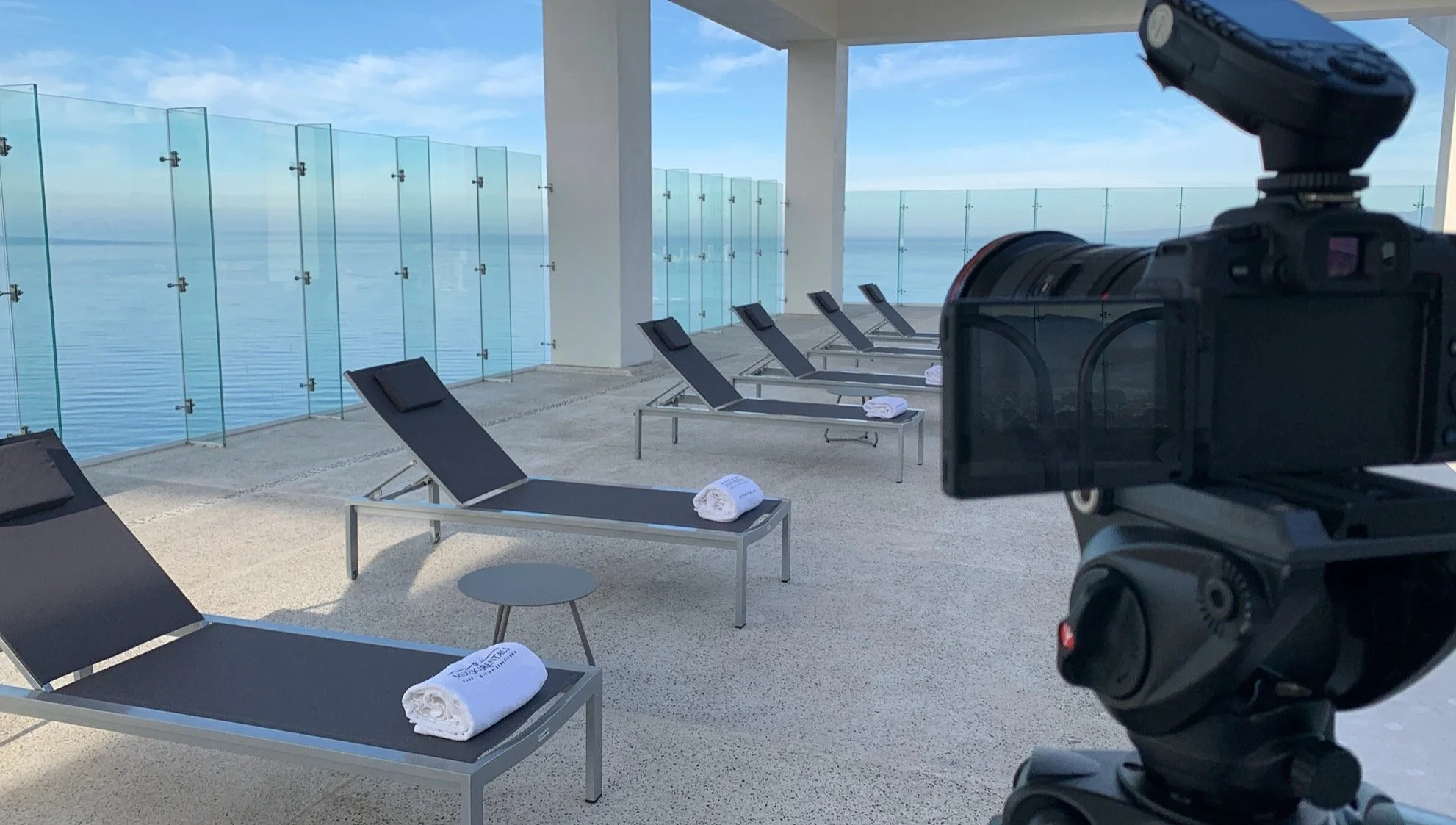 Rooftop lounge area with black lounge chairs and rolled white towels, surrounded by glass railings overlooking the ocean of Puerto Vallarta, with a camera in the foreground.