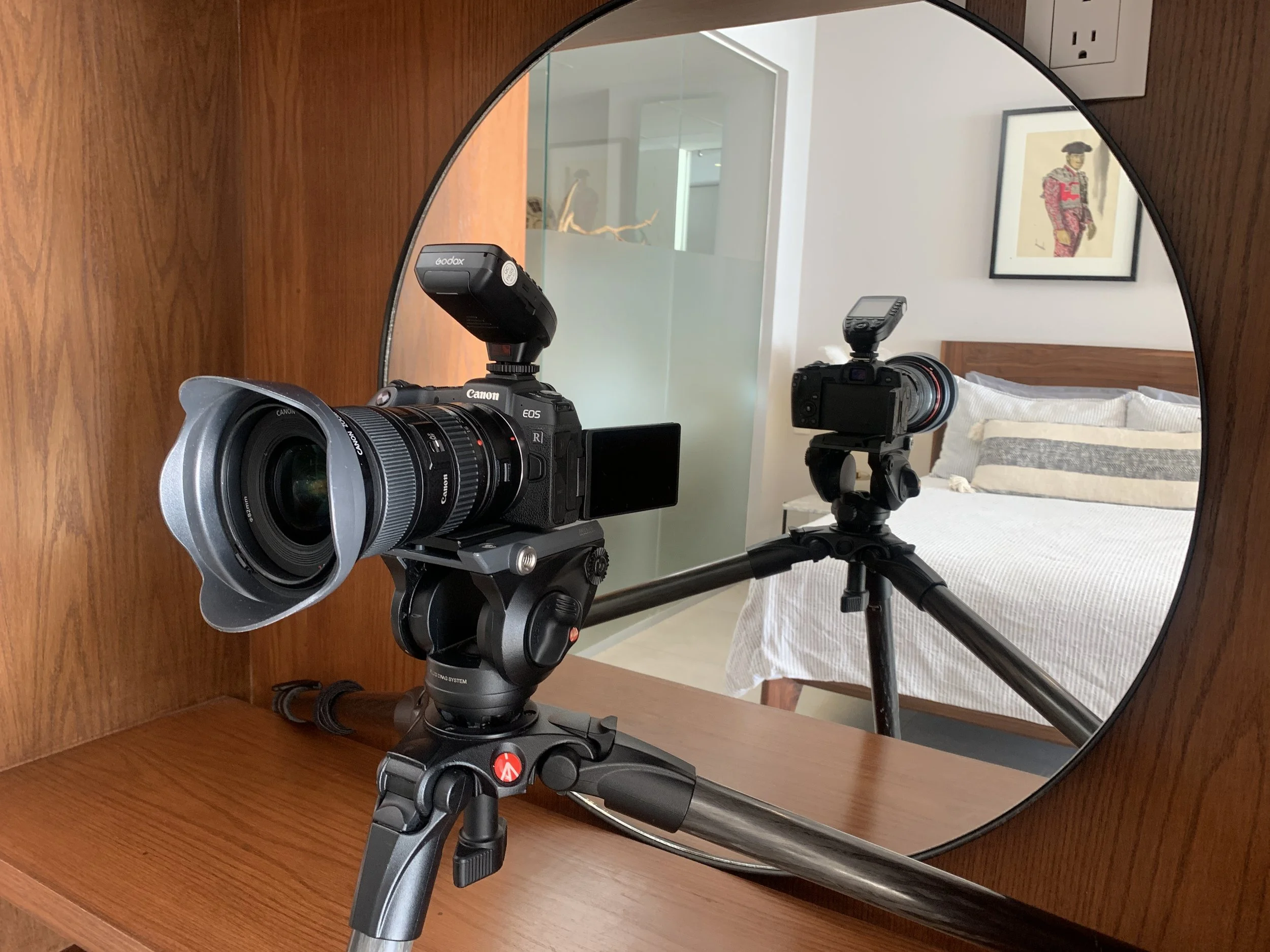 A mirror reflects a camera with a telephoto lens mounted on a tripod, positioned on a wooden surface, with a bedroom and framed artwork visible in the background.